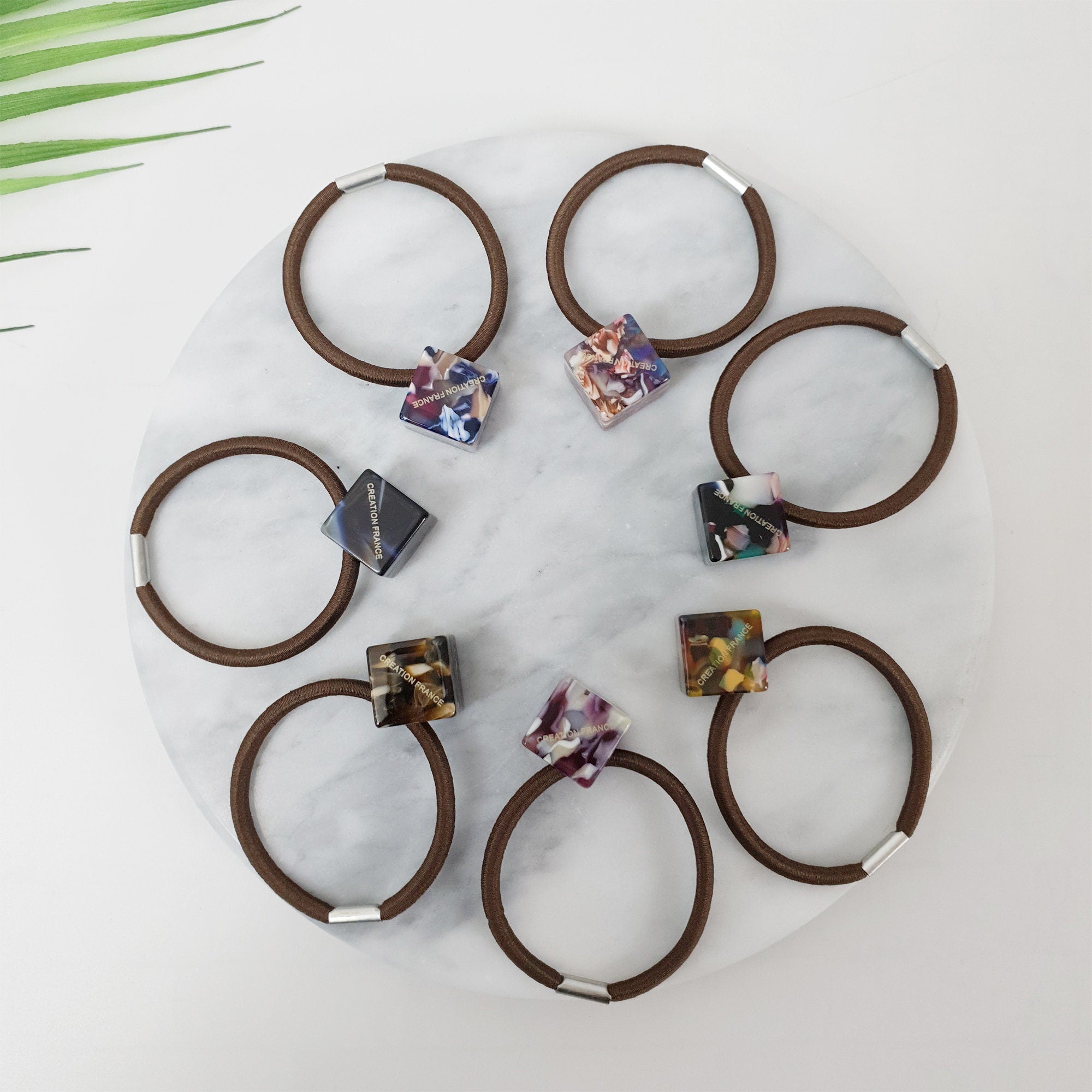 Cellulose acetate cube hair tie in tortoise shell and marble patterns. Geometric square ponytail holder. Modern Korean hair accessory. Six brown hair ties with square shaped charms. The charms are made of clear plastic with a colorful marbled pattern and gold lettering that reads 'CREATION GRACE'. The charms are attached to the hair ties with a silver metal ring.