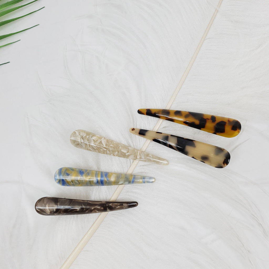 Cellulose acetate alligator hair clip in tortoise and marble patterns featuring a sleek beak design for side bangs and half-up styling. Five hair clips with different patterns. The clips are made of tortoiseshell, brown, and white plastic. The clips are arranged in a row on a white feathery surface.