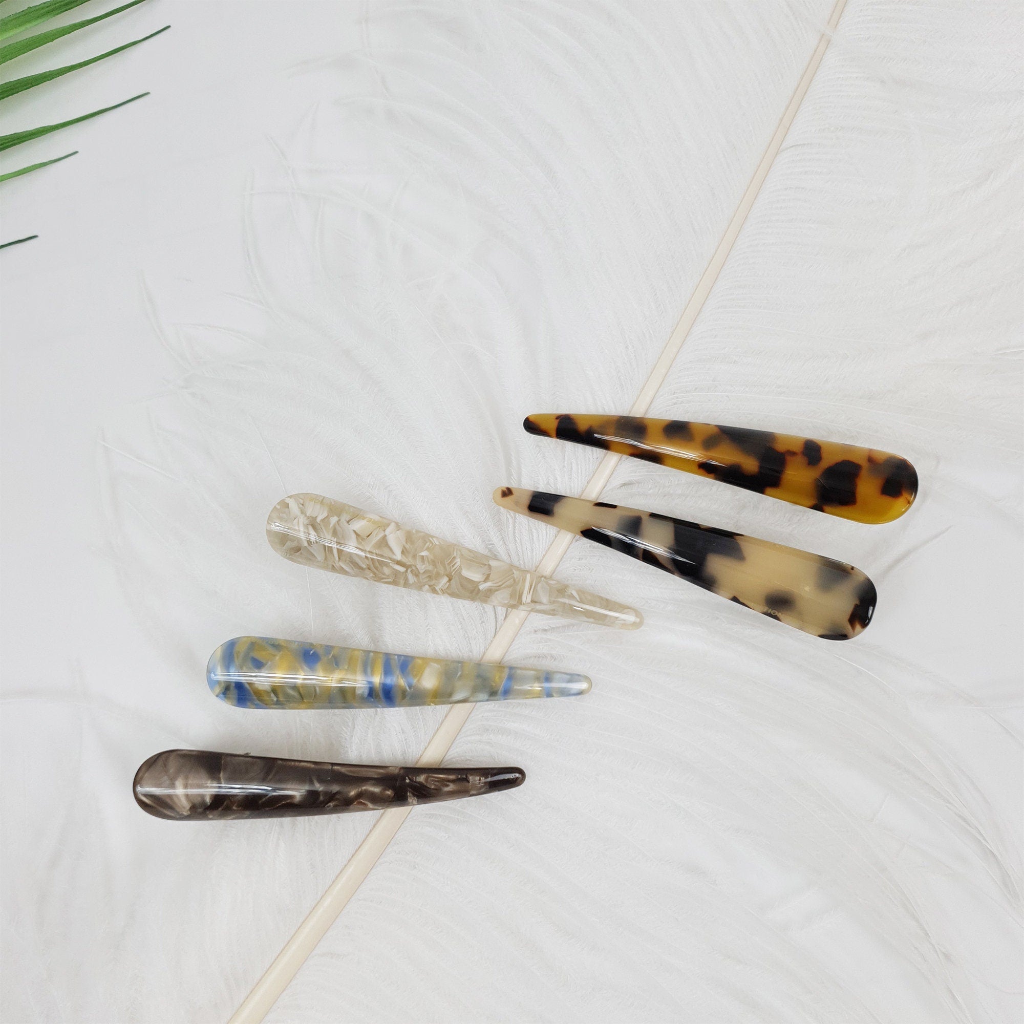 Cellulose acetate alligator hair clip in tortoise and marble patterns featuring a sleek beak design for side bangs and half-up styling. Five hair clips with different patterns. The clips are made of tortoiseshell, brown, and white plastic. The clips are arranged in a row on a white feathery surface.
