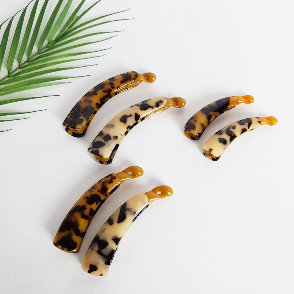 Three sizes of cellulose acetate banana clips in tortoise shell patterns, interlocking comb teeth for secure ponytails and updos, classic retro hair styling tool. Six tortoise shell and white hair clips in various sizes. The clips are curved and have a mottled pattern.