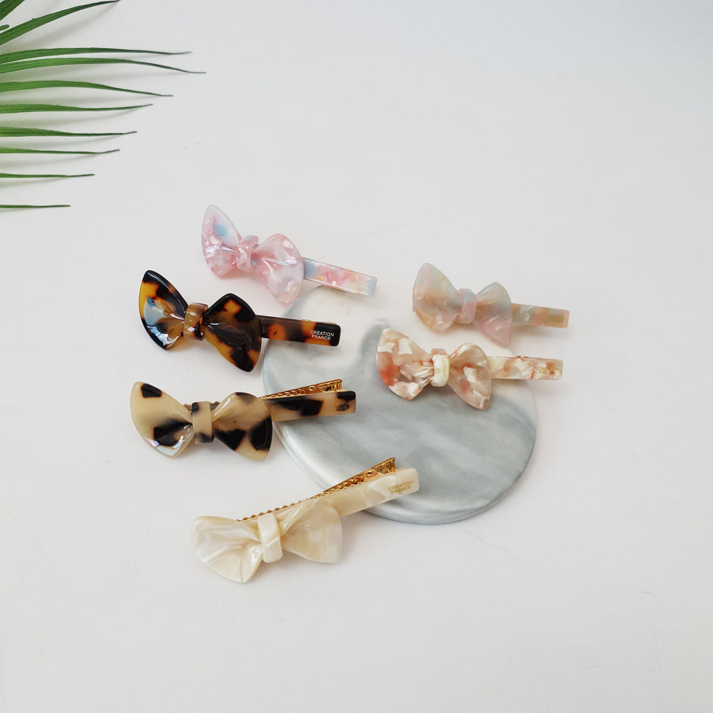 Cellulose acetate ribbon hair clip with gold alligator beak clasp. Small glossy bow barrette in pink marble and tortoise shell patterns for styling bangs or side hair.