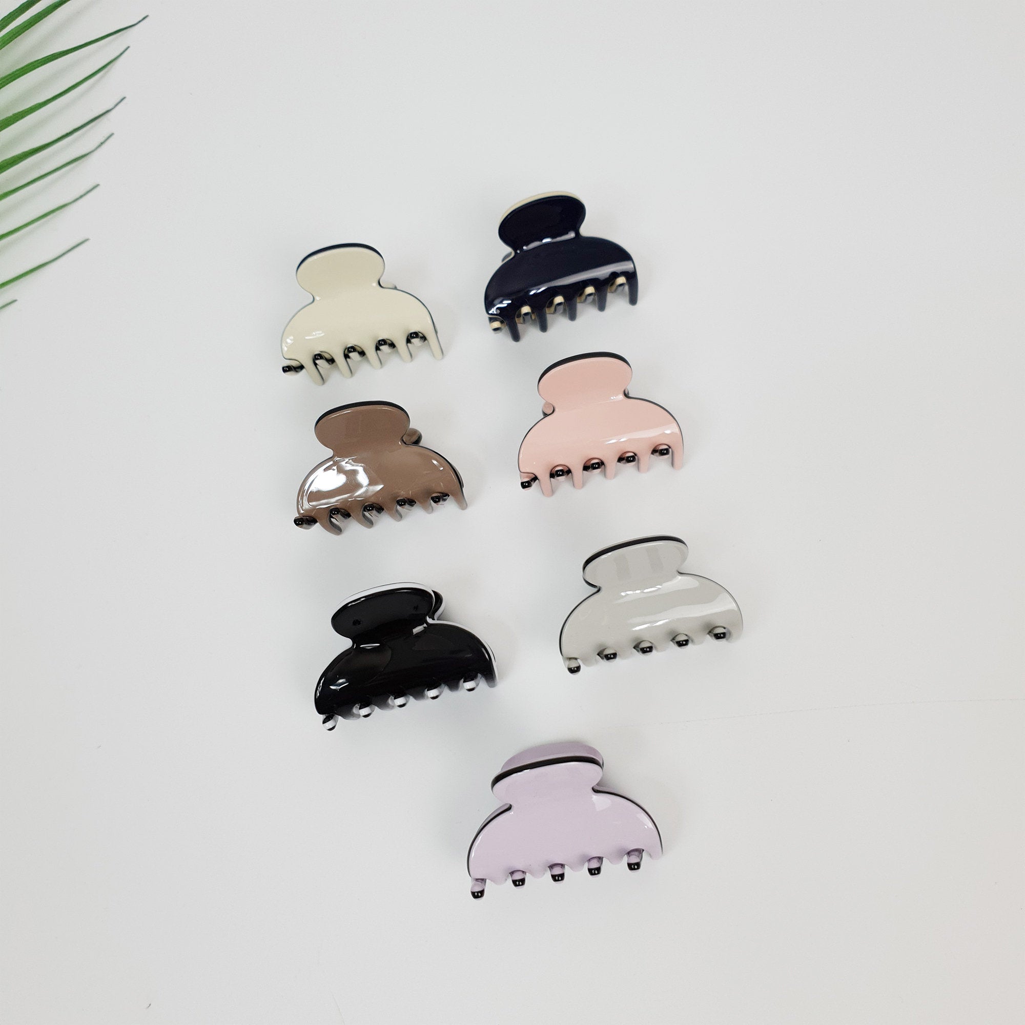 Mini acetate hair claw clips in pastel colors. Small half moon jaw clips for styling bangs or side hair. Tiny decorative hair accessories for women and toddlers.  Seven plastic hair clips in various colors including black, white, brown, pink, and light purple. The clips are all the same shape and size, with a rounded top and four prongs on the bottom.