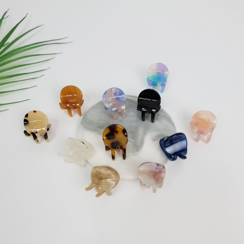 Glossy cellulose acetate mini hair claw clips in bean shape. Small 2cm tortoise shell and solid color accessories for holding side bangs. Cute minimal hair jewelry for girls and women. A collection of twelve small, colorful, plastic hair clips. The clips are arranged in a circle on a gray and white marble surface. The clips are in various colors, including brown, black, white, blue, pink, and purple. The clips are all in the shape of a half-circle with two prongs on the bottom.
