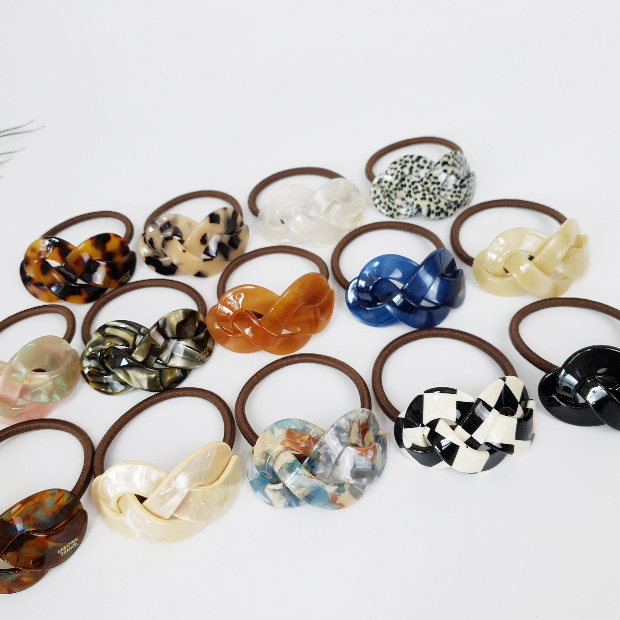 Cellulose acetate twisted hair tie in tortoise shell, marble, and checkered patterns. Geometric knot ponytail holder for women with a curved design.  Assortment of brown, black, white, blue, and multi-colored hair ties with a knotted design. The hair ties are made of plastic and have a glossy finish.