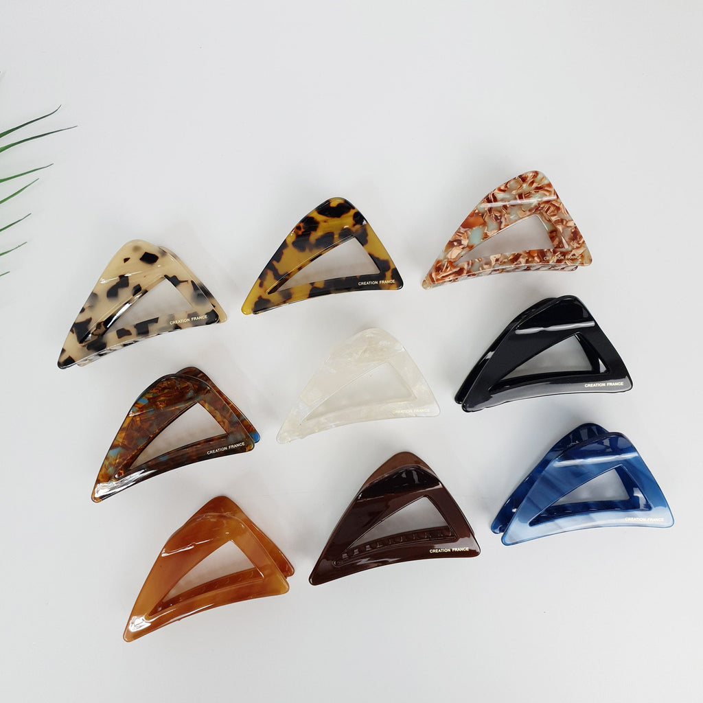 Cellulose acetate triangle hair claw clip in tortoise shell and solid colors. 3.0 inch geometric jaw clamp for modern half-up hairstyles. Assortment of eight triangle shaped hair clips in various colors and patterns including brown, black, white, blue, and tortoise shell.  Each clip has a small gold label that reads 'CREATOR FRANCE'.