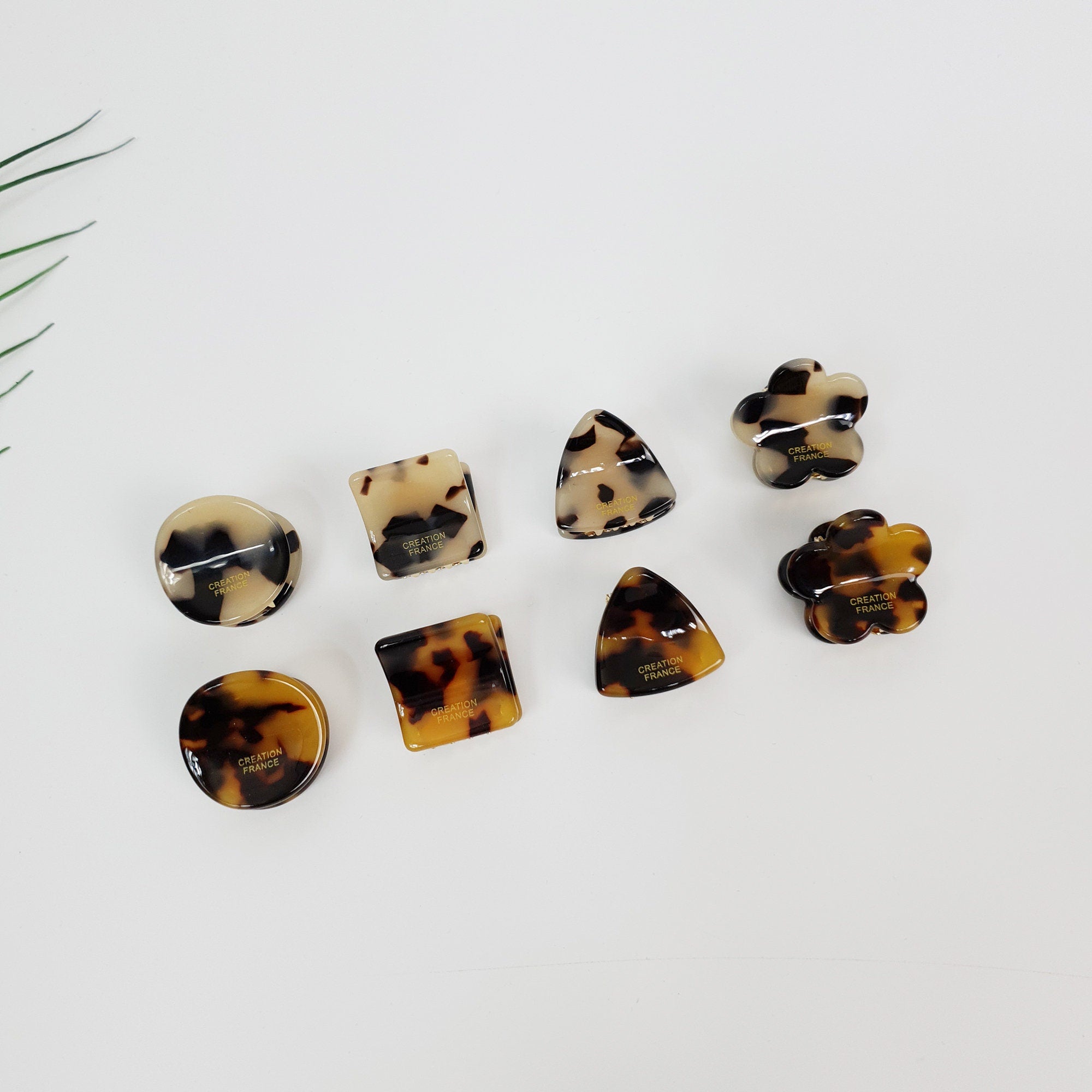 Cellulose acetate tortoise shell mini hair claw clips in geometric shapes including square, triangle, and flower. Brown and beige leopard pattern small jaw clips for women. Minimalist Korean style hair accessories for bangs and half-up hairstyles.  Eight tortoise shell hair clips in various shapes including a square, a circle, a flower, a triangle, and a butterfly. The clips are all brown and black with a gold inscription that reads 'CREATION FRANCE'.