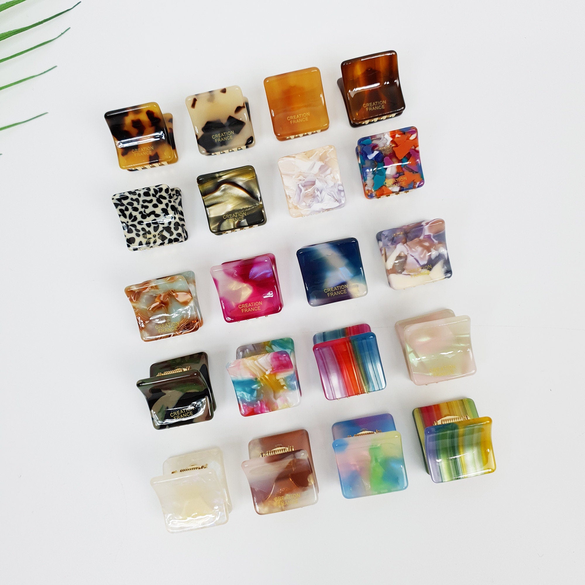 Miniature square cellulose acetate hair claw in tortoise and art patterns, 0.8 inch geometric cube shape for bangs, non-slip tiny jaw clip for side hair styling. A collection of 18 square hair clips with various colorful patterns and designs. The clips are made of plastic and have a gold metal clasp. The clips are arranged in rows on a white surface. The clips have the words 'Creation France' printed on them.