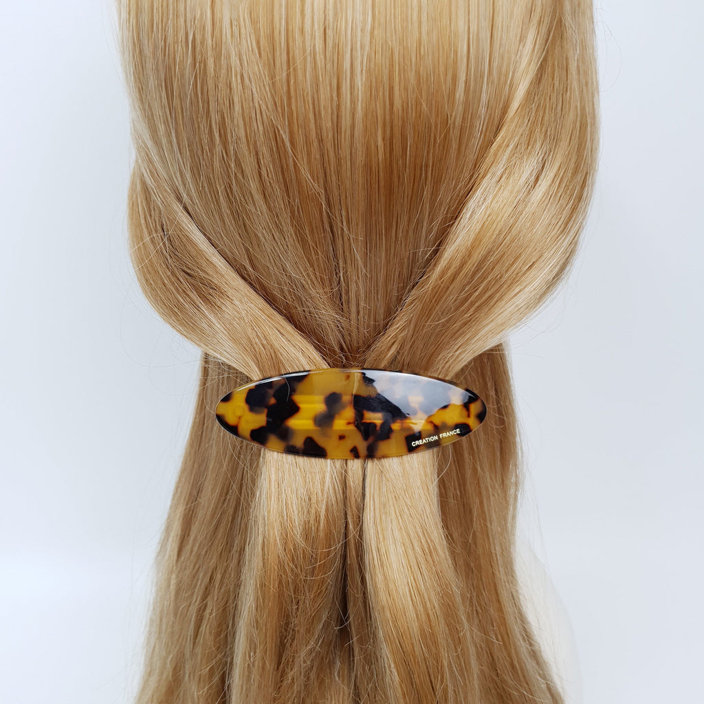 Oval cellulose acetate hair barrette in tortoise shell and solid colors, flat smooth finish with automatic metal clasp, minimalist hair accessory for half-up styles. A brown and black tortoise shell hair clip with the words 'CREATION FRANCE' printed on the back.