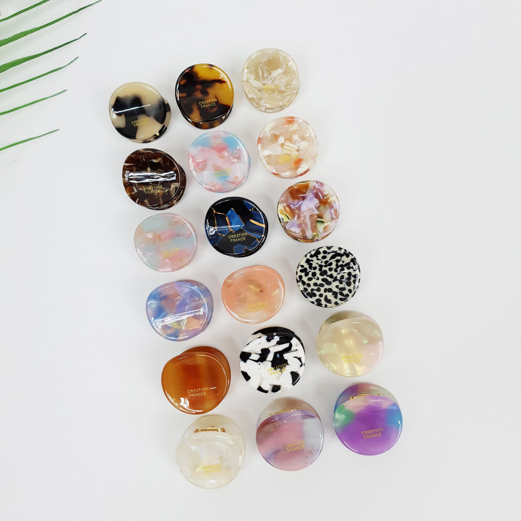 Cellulose acetate mini hair claw clips in tortoise shell and marble patterns, round small french pin for bangs and side hair styling, cute non-slip accessories for women and girls. A collection of 17 round hair clips in various colors and patterns, including black and white, brown and white, pink and white, blue and white, and multi-colored. The clips are made of plastic and have a smooth, glossy finish. Some of the clips have the words 'Creation France' printed on them.