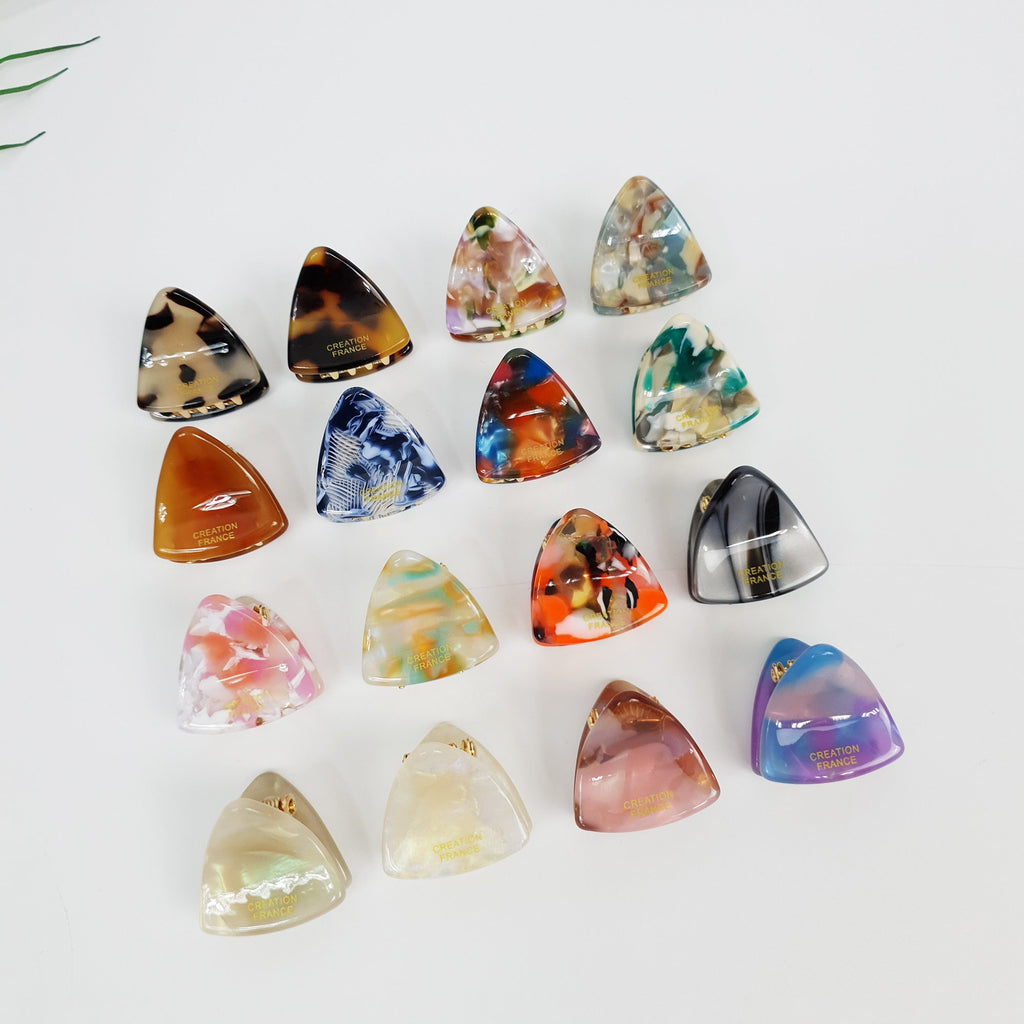 Cellulose acetate triangle mini hair claw clips in tortoise shell and marble patterns. Small geometric jaw clamps with gold spring for women's bangs and side hair styling. Minimalist Korean fashion accessory. A collection of 15 triangular hair clips with various patterns and colors. The clips are made of plastic and have a gold inscription that reads 'CREATION FRANCE'.