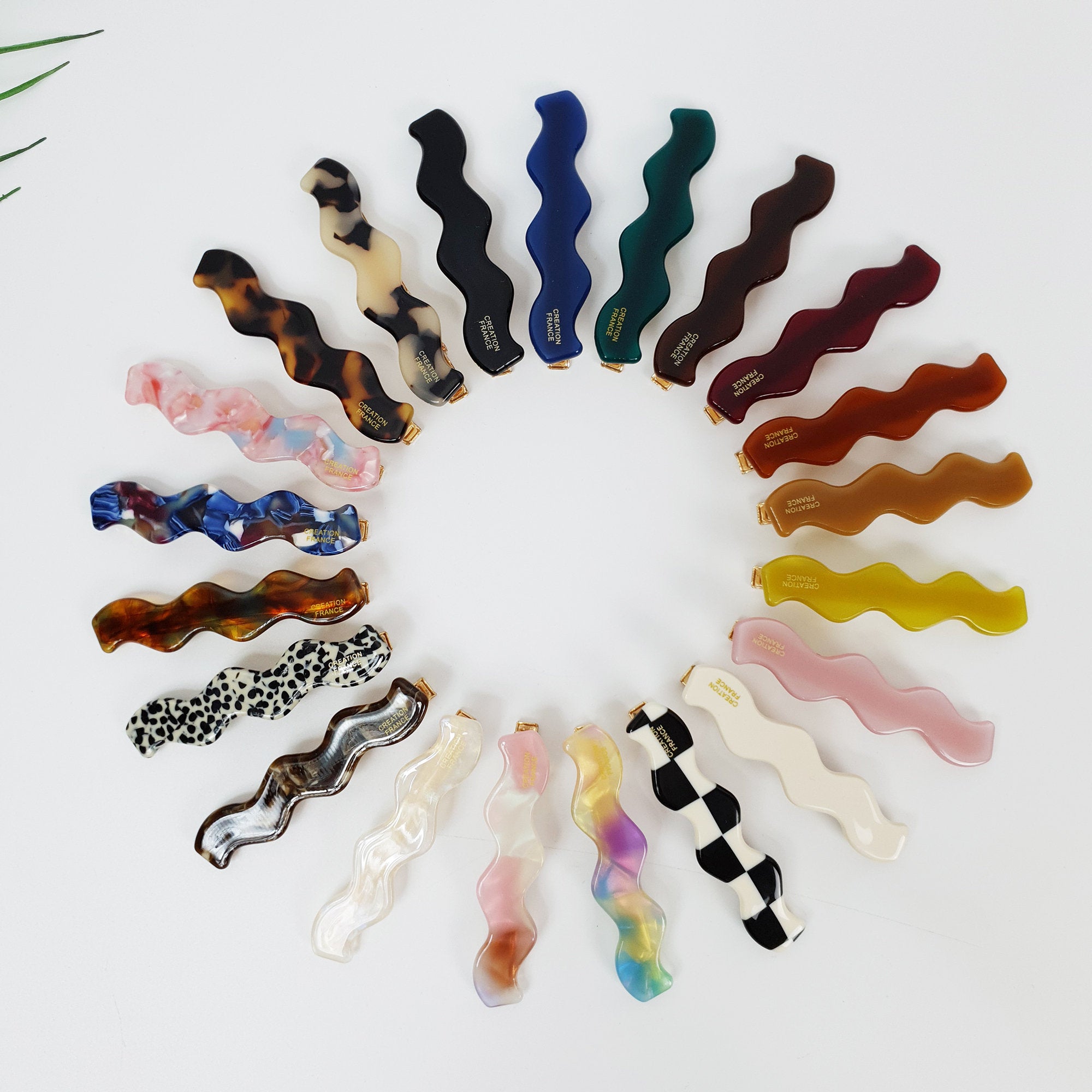 Wavy cellulose acetate hair barrette in tortoise and pastel colors, 2.6 inch squiggle shape alligator clamp for side bangs, trendy minimalist styling tool. A collection of 18 wavy hair clips in various colors and patterns including brown, black, white, pink, blue, green, yellow, and leopard print. The clips are arranged in a circle on a white background.