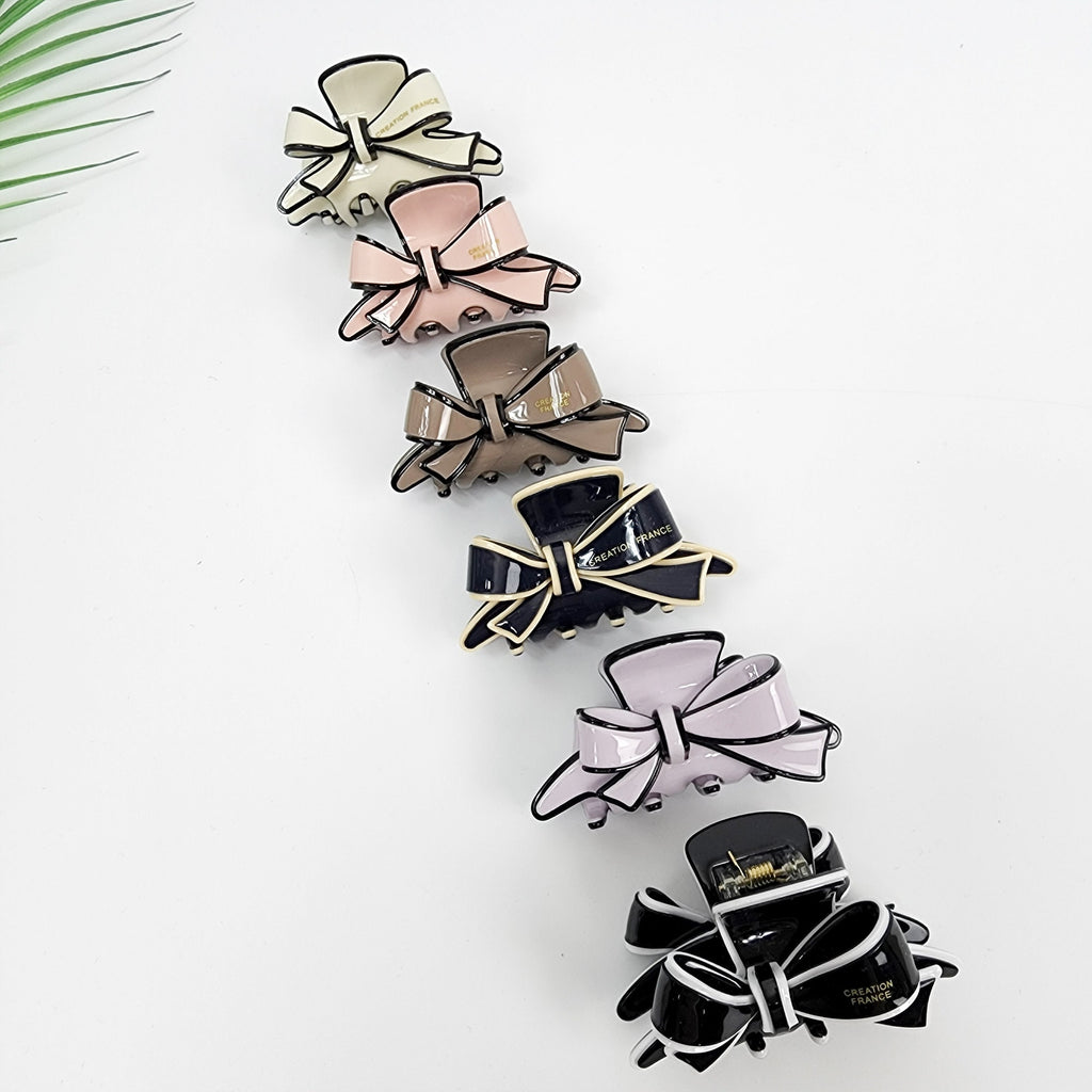 Pastel cellulose acetate asymmetrical ribbon hair claw clip. 2.4 inch fashion bow jaw clamp. Elegant hair accessory for half updos. A collection of six bow-shaped hair clips in various colors. The clips feature a glossy finish with a contrasting black trim. Colors include cream, pink, brown, black, and lavender. Each clip has the words 'CREATION FRANCE' printed on it.