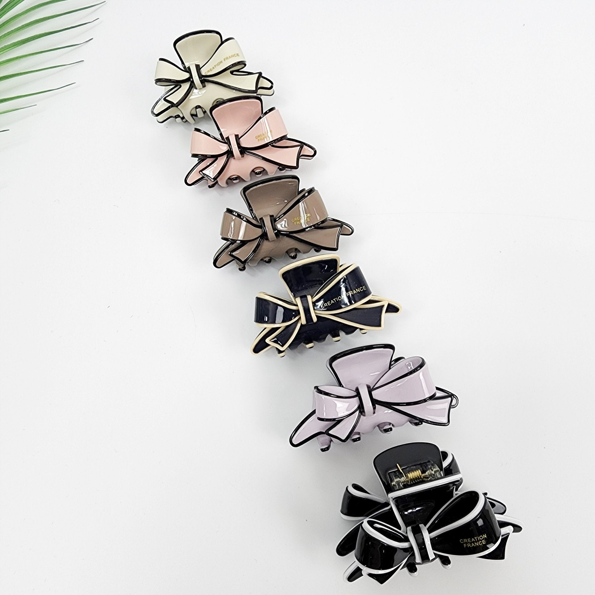 Pastel cellulose acetate asymmetrical ribbon hair claw clip. 2.4 inch fashion bow jaw clamp. Elegant hair accessory for half updos. A collection of six bow-shaped hair clips in various colors. The clips feature a glossy finish with a contrasting black trim. Colors include cream, pink, brown, black, and lavender. Each clip has the words 'CREATION FRANCE' printed on it.