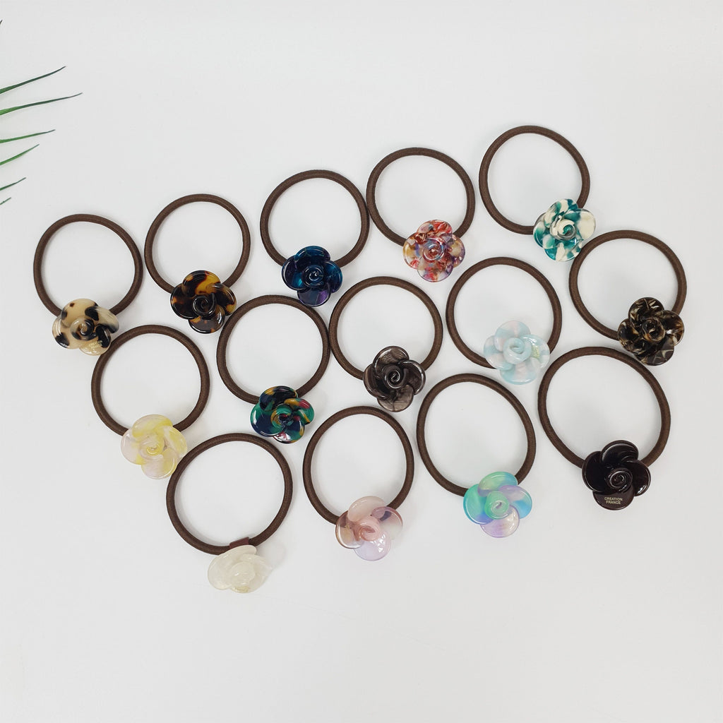 Acetate flower hair tie with camellia design. Tortoise shell and marble pattern rose ponytail holder. Cute Korean hair accessory for women.  A collection of brown hair ties with various colored flower embellishments. The flowers are made of plastic and have a variety of patterns and colors, including blue, green, pink, purple, and brown.