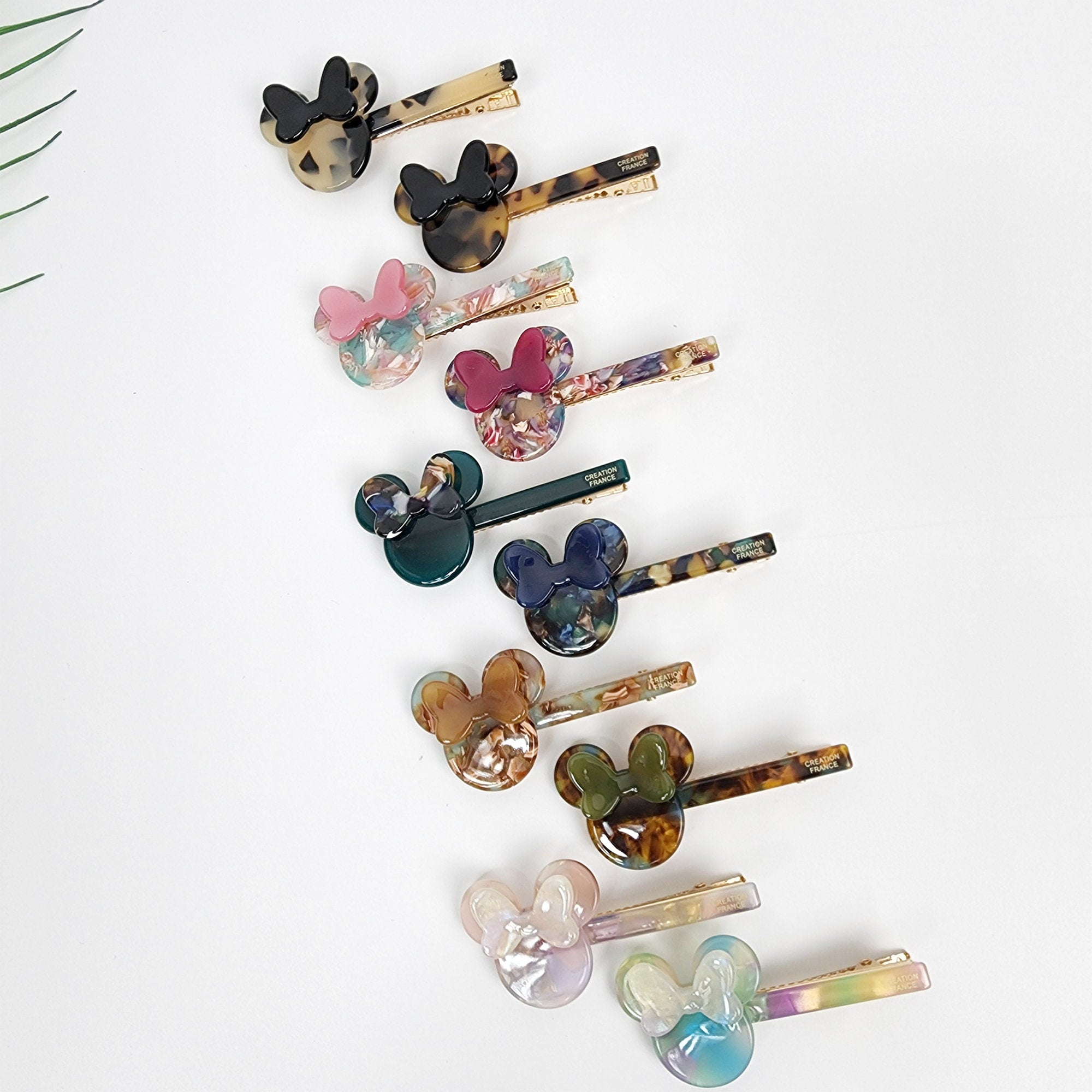 Small cellulose acetate mouse ear hair clip with bow. Tortoise shell and pastel marble alligator clamp. 2.7 inch cute side pin for bangs and girls. Assortment of colorful hair clips with a cartoon mouse design. The clips are made of plastic and have a gold metal clip. The clips are arranged in a row on a white background.
