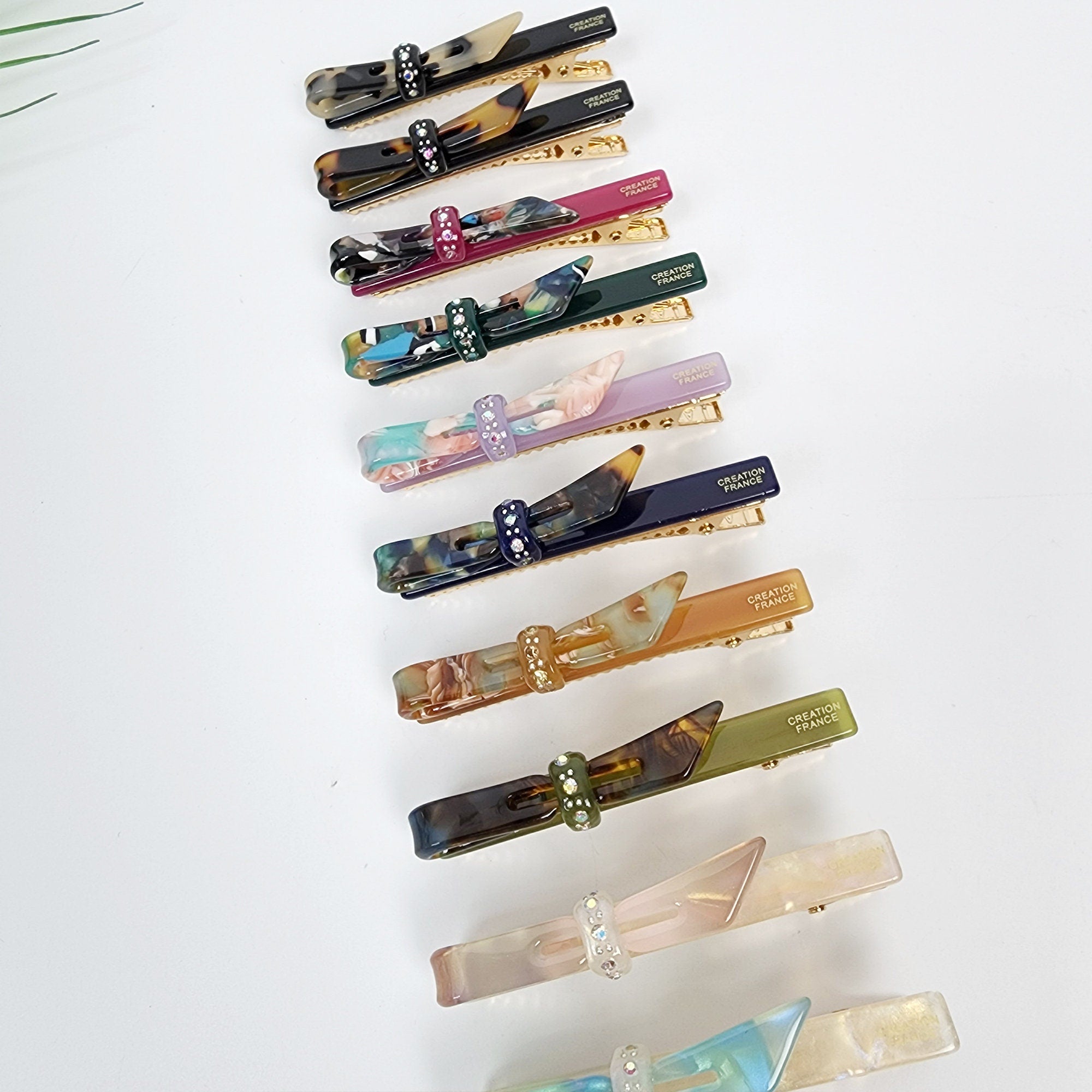 Small cellulose acetate bow hair clip with rhinestone detail. Tortoise shell and colorful alligator clips for bangs. 2.6 inch side hair pin for women and girls. A set of twelve hair clips with bows. The bows are made of different colored and patterned plastic. Each bow has a rhinestone in the center. The clips are gold colored and have the words 'CREATION FRANCE' printed on them.