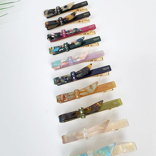 Small cellulose acetate bow hair clip with rhinestone detail. Tortoise shell and colorful alligator clips for bangs. 2.6 inch side hair pin for women and girls. A set of twelve hair clips with bows. The bows are made of different colored and patterned plastic. Each bow has a rhinestone in the center. The clips are gold colored and have the words 'CREATION FRANCE' printed on them.
