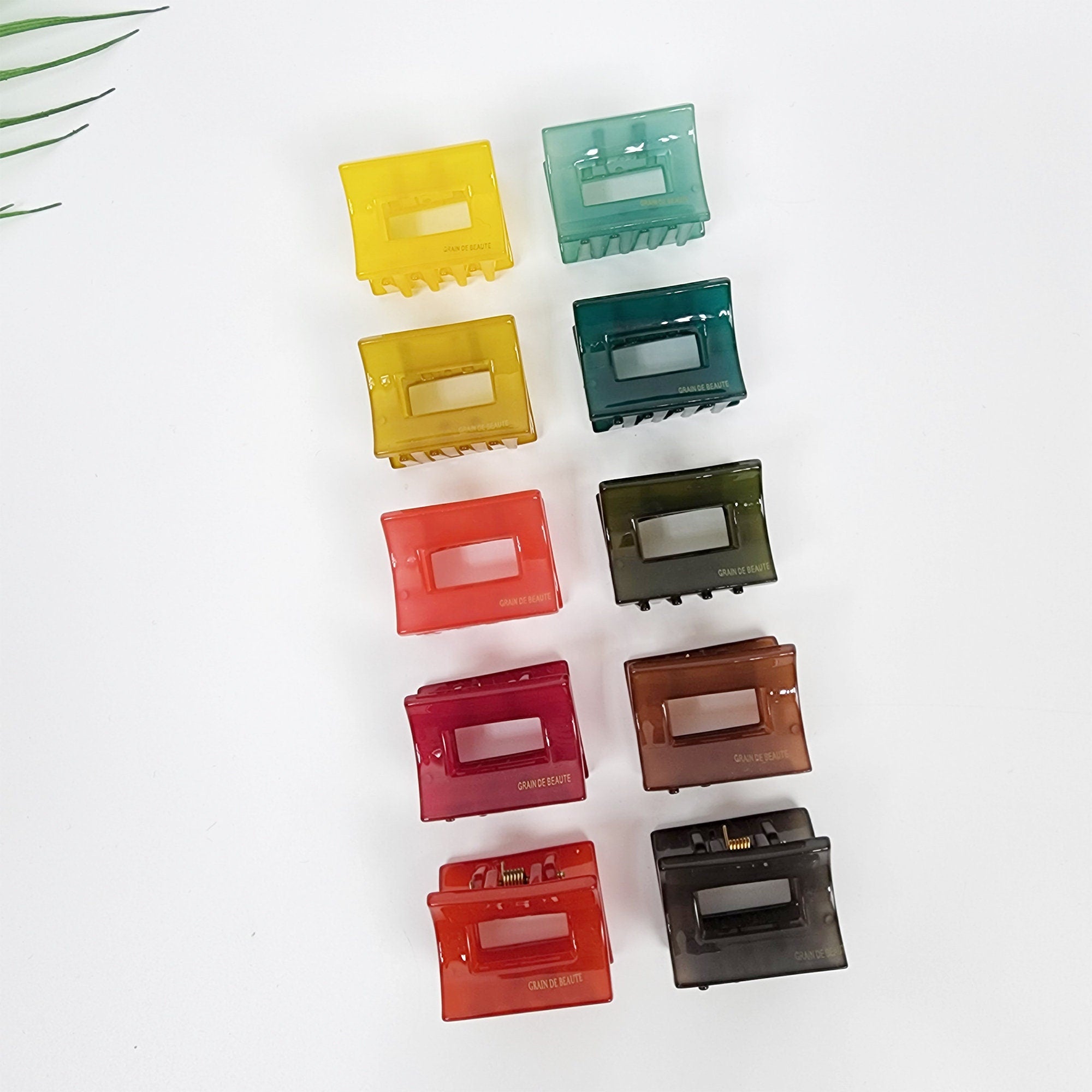 Small square cellulose acetate hair claw clip. Translucent solid color geometric jaw clamp. 1.5 inch mini accessory for half updos and bangs. Ten colorful plastic hair clips arranged vertically. The clips are in various shades of green, yellow, red, and brown. The clips are all rectangular with a rectangular opening in the center. The clips have the words 'Grain de Beauté' printed on them.
