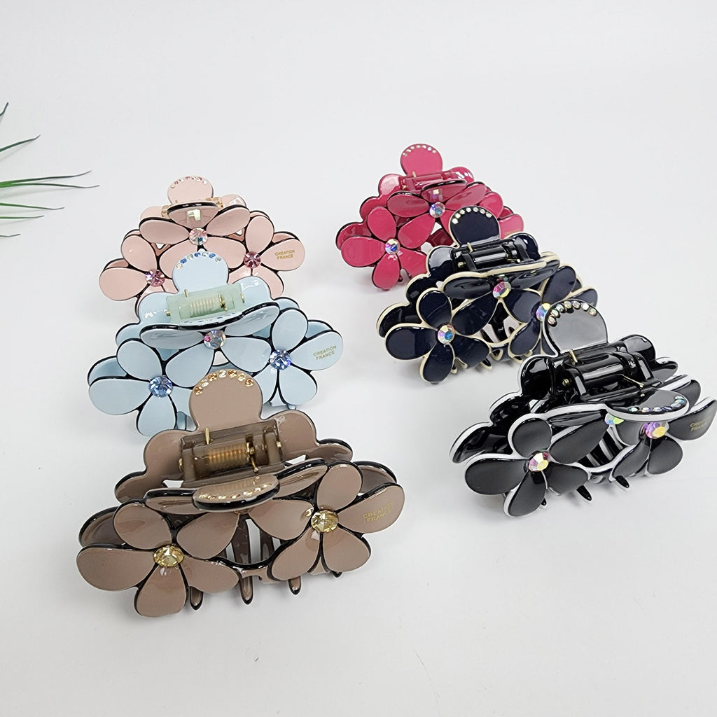 Cellulose acetate flower hair claw with rhinestones. Medium floral jaw clip for half-up hairstyles available in pastel and dark colors.  Six flower-shaped hair clips in various colors including pink, red, blue, and black. Each clip has a flower design with rhinestones and a small metal clasp. The clips are arranged on a white surface.
