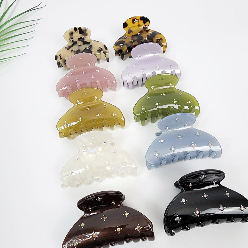 Cellulose acetate hair claw clip with rhinestone star details, medium non-slip jaw clip for half-up hairstyles or thick hair accents. Eight hair clips with rhinestones. The clips are in various colors including brown, green, light blue, dark blue, pink, white, and black. The clips are all the same shape and size, with a curved, claw-like design.
