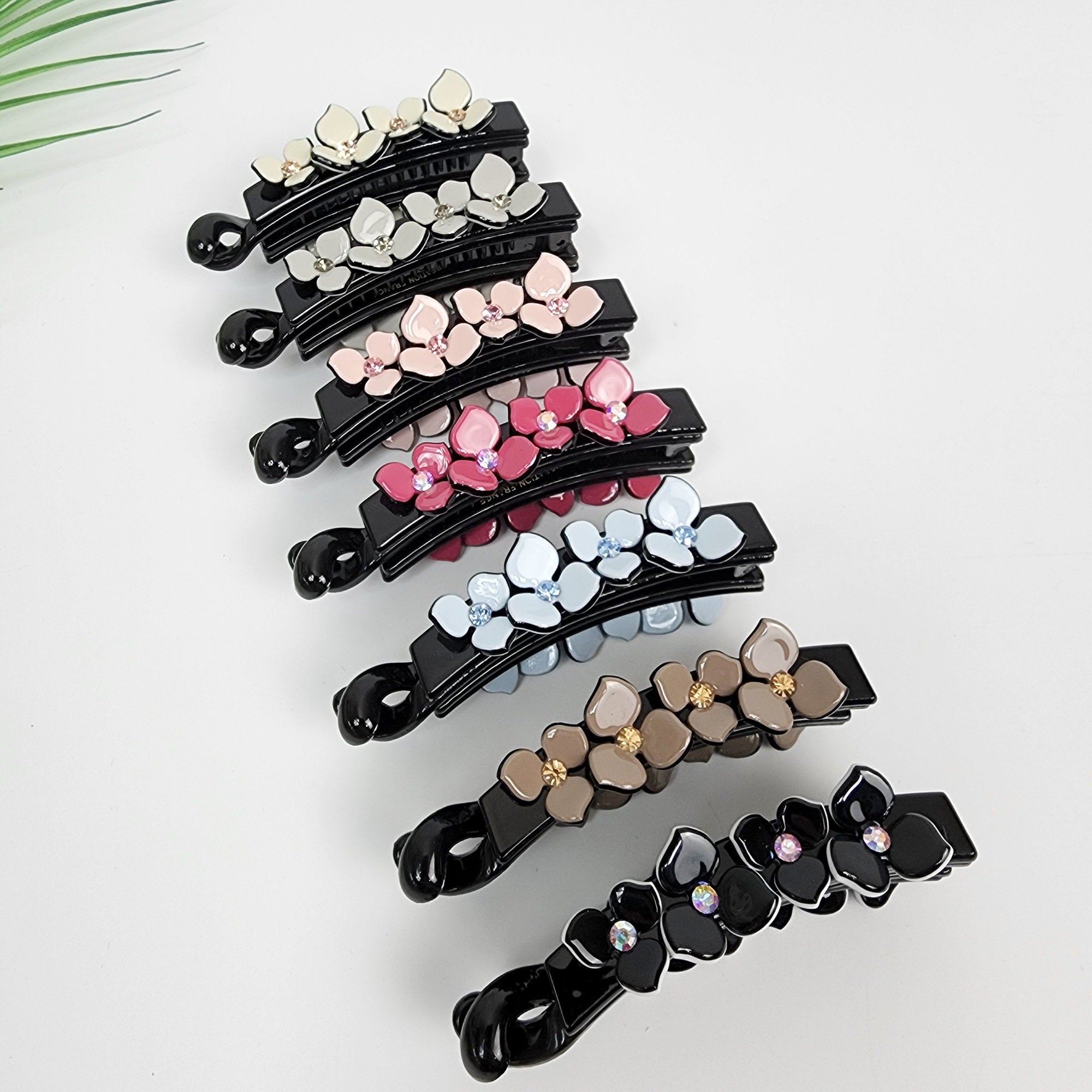 Small floral banana clip with rhinestone crystals and interlocking teeth, non-slip acetate hair accessory for thin hair or half-up styles. Seven black hair clips with different colored flower designs. The clips are arranged in a stack with the largest clip at the bottom and the smallest clip at the top. The flower designs are in shades of pink, blue, and white.