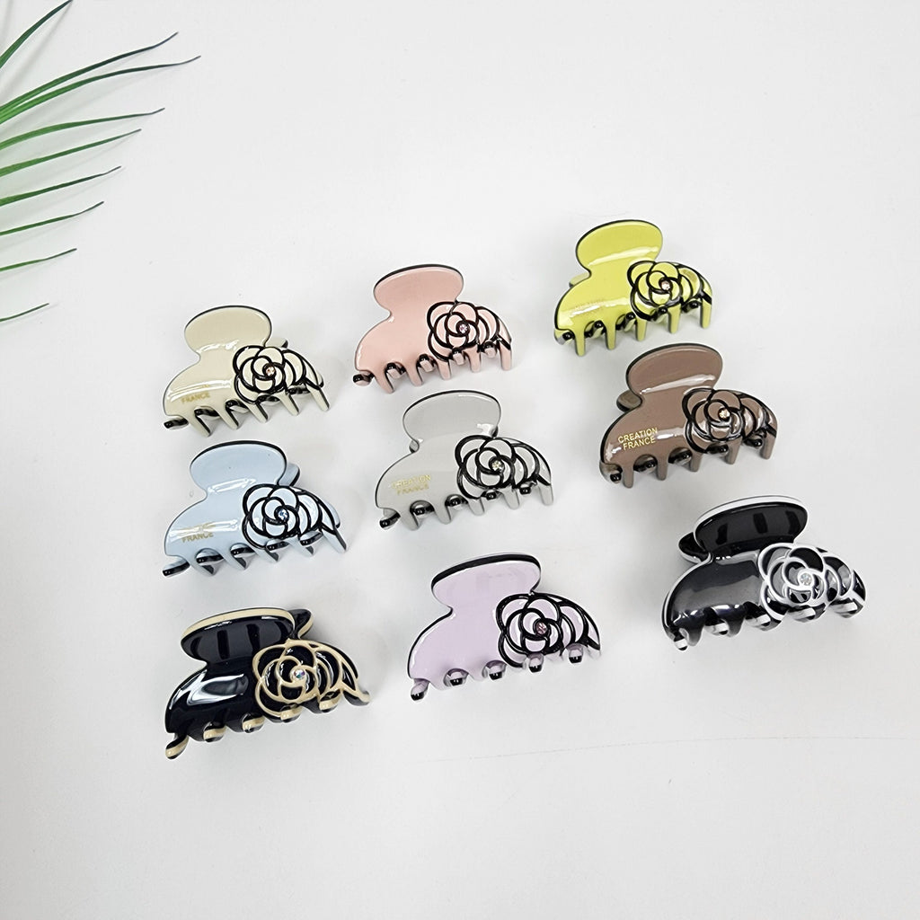 Mini cellulose acetate hair claw featuring a camellia flower design with black outline and crystal center. Pastel round jaw clip for side bangs styling. Nine hair clips with a flower design. The clips are in various colors including pink, yellow, light blue, gray, brown, and black. The clips have the words 'Creation France' printed on them.