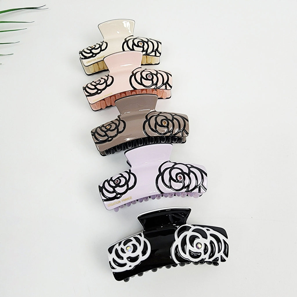Cellulose acetate hair claw clip with camellia flower pattern and rhinestone center, medium double tooth clamp for secure half up styling. Five hair clips with black floral designs on a white background. The clips are stacked on top of each other. The top clip is beige, the next is pink, the next is brown, the next is lavender, and the bottom clip is black.