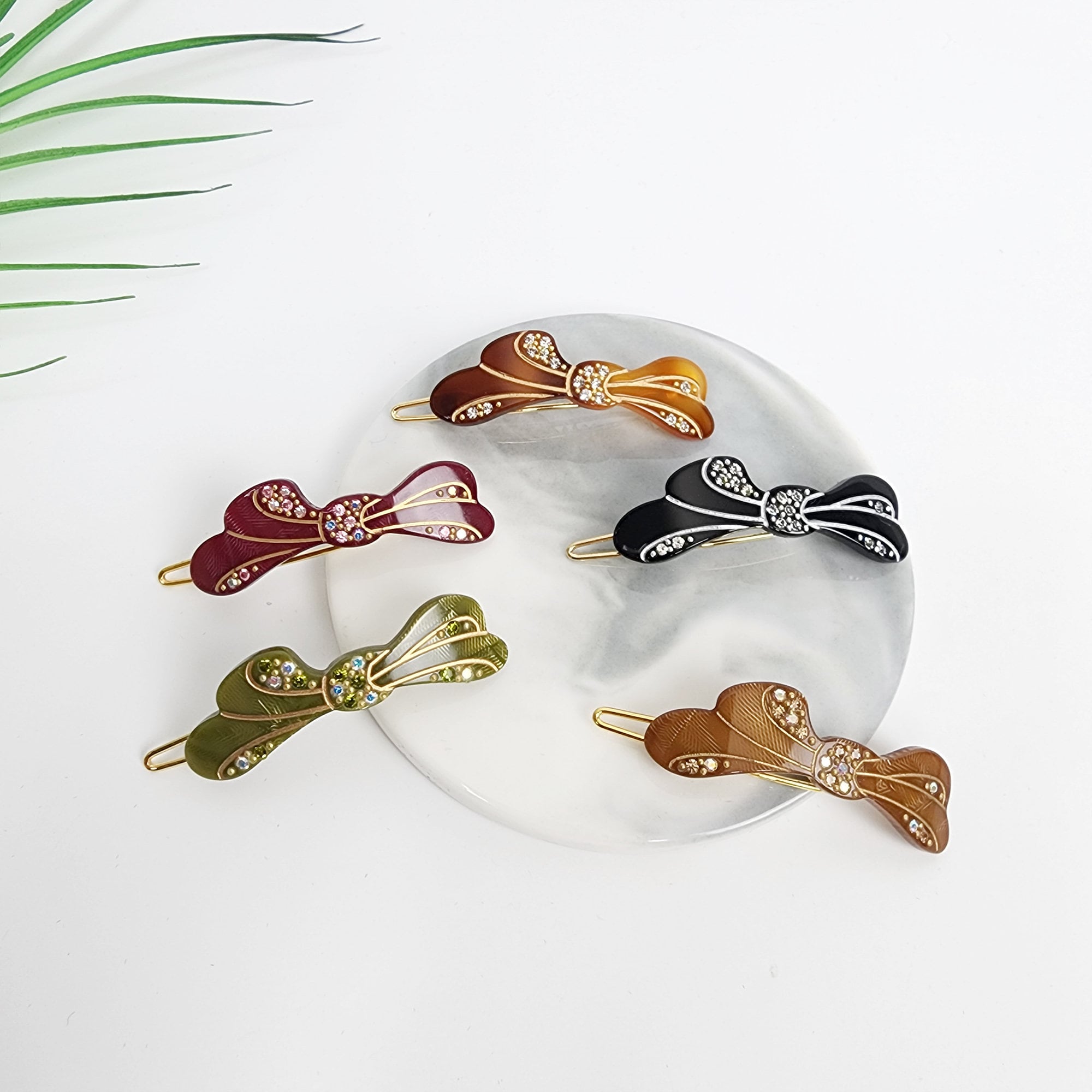 Sparkly cellulose acetate bow hair clip with rhinestone accents, small snap barrette for bangs and side hair styling.Five hair clips with jeweled bows in different colors: green, red, black, brown, and gold. The clips are arranged on a white marble surface.