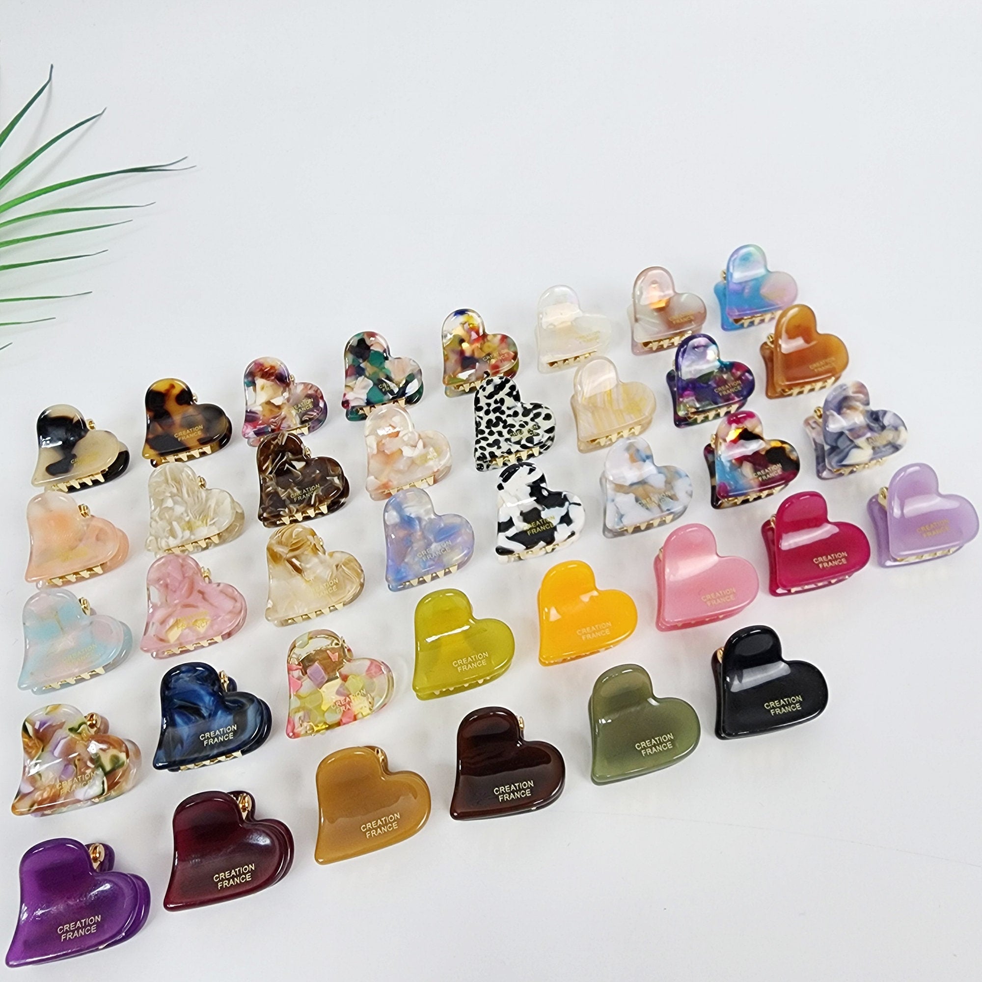 Cellulose acetate heart shape mini hair claw clips in tortoise shell and marble patterns. Small geometric jaw clamps with gold spring for women's bangs and side hair styling. Cute Valentine gift accessory. A collection of 30 heart-shaped hair clips in various colors and patterns. The clips are made of plastic and have a gold metal clasp. Some of the clips have a 'Creation France' logo on them. The clips are arranged in a semi-circle on a white surface.