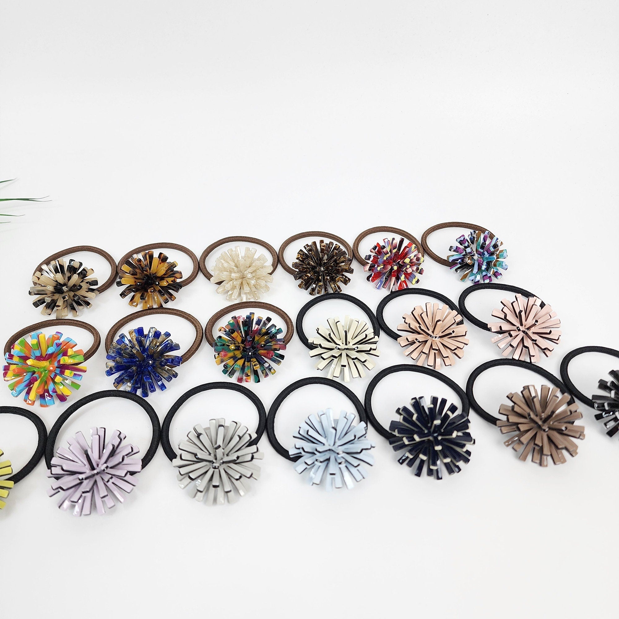 Cellulose acetate dandelion hair tie in tortoise shell and pastel colors. Spiky 3D pom pom ponytail holder. Glossy Korean hair accessory for women. A collection of 20 hair scrunchies with different colored and patterned fabric flowers. The scrunchies are arranged in rows on a white surface.