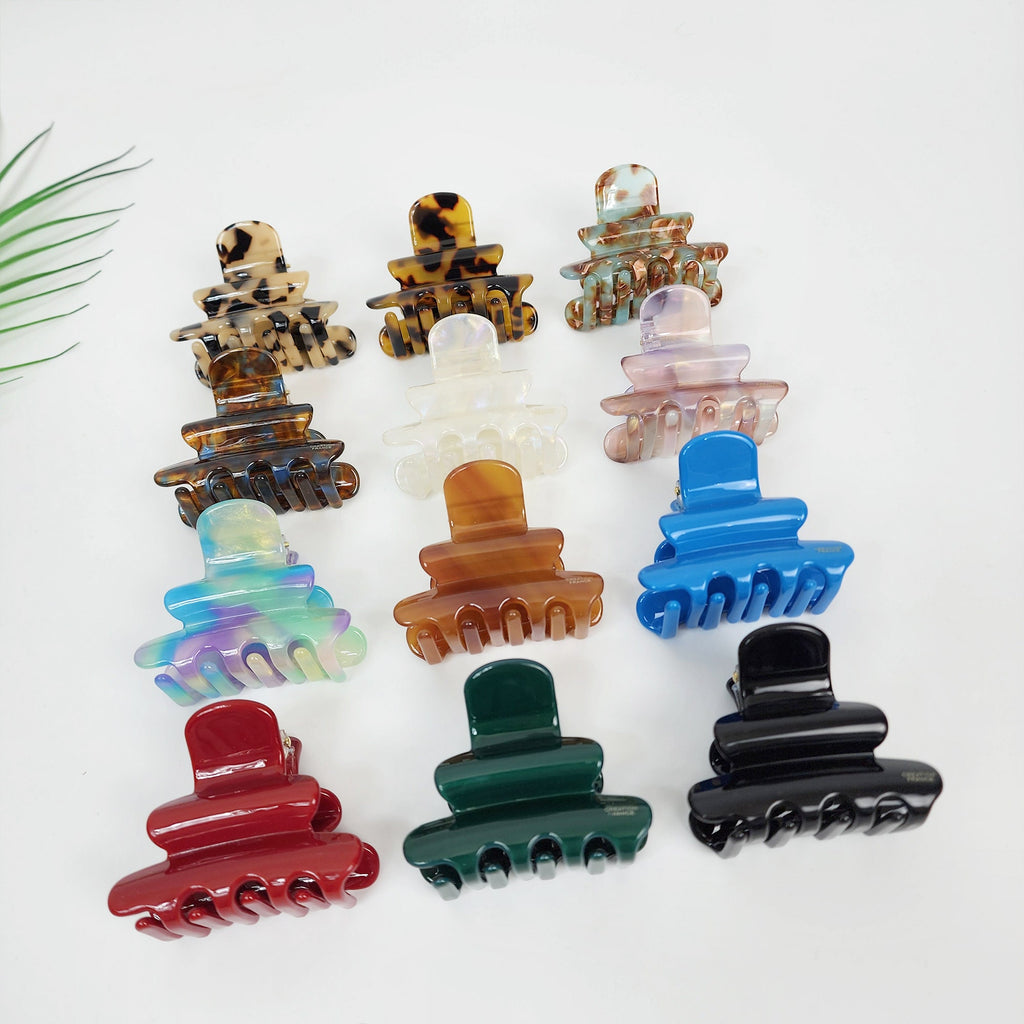 Cellulose acetate hair claw clip in tiered bell shape featuring tortoise shell and aurora patterns for women half up hairstyle and daily accessory. Twelve assorted plastic hair clips in various colors, including brown, white, blue, green, red, and iridescent. The clips are all the same size and shape, with a curved top and four prongs on the bottom.