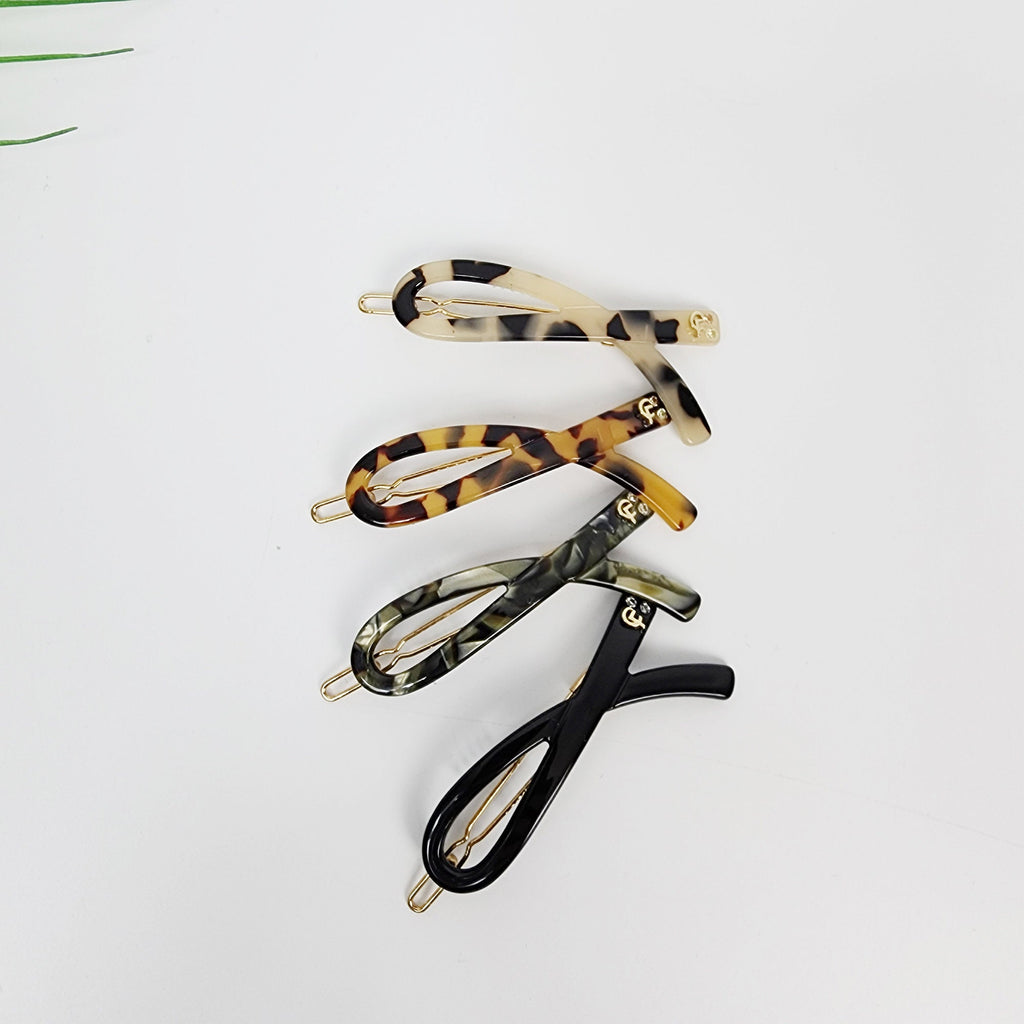Tortoise shell acetate hair clip in twisted scarf shape, minimalist snap barrette for bangs and side hair styling. Four hair clips in different patterns. The clips are all made of a tortoiseshell-like material and have a gold accent. The clips are arranged in a stack, with the top clip being a light brown and white pattern, the second clip being a brown and black pattern, the third clip being a green and black pattern, and the bottom clip being a black pattern.