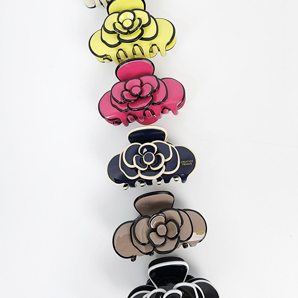 Cellulose acetate flower hair claw in black and white with gold outline. Small 2.4 inch camellia jaw clip for half-up hairstyles and feminine styling. Five hair clips with a flower design. The clips are in different colors: yellow, pink, blue, brown, and black. The clips have a black outline and a gold inscription that reads 'Creation France'.