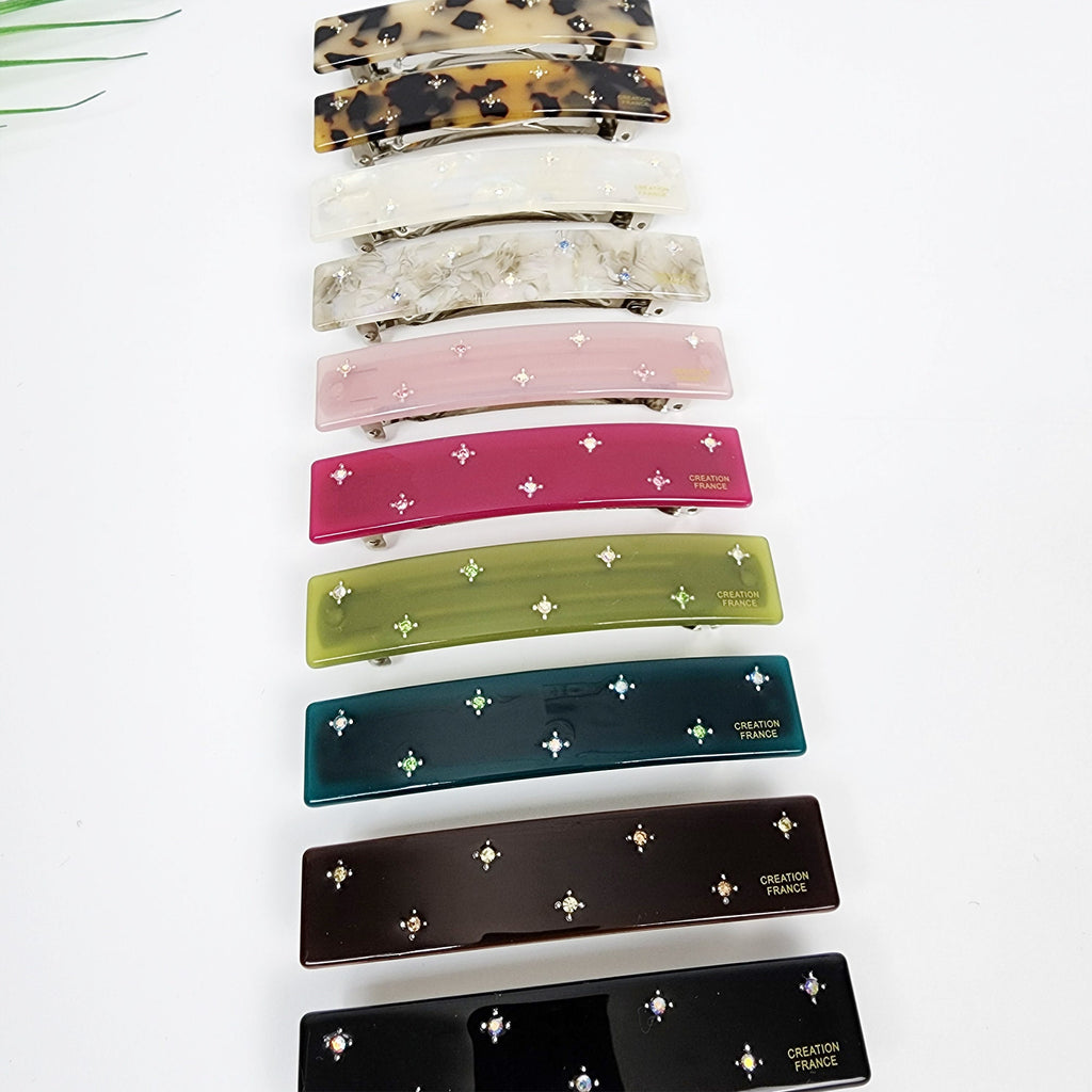 Cellulose acetate rectangular barrette featuring star cubic accents and french clip mechanism for women half up hairstyle and daily accessory. Ten hair clips with a variety of colors and designs. The clips are all made of plastic and have a shiny finish. The clips have a curved shape and are decorated with small, clear rhinestones. The clips have the words 'CREATION FRANCE' printed on them. The colors of the clips are: brown, white, gray, pink, red, green, teal, brown, black, and black.