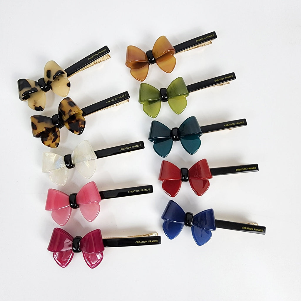 Cellulose acetate bow hair clip with alligator grip in tortoise shell pattern. 3.5 inch preppy ribbon barrette for women holding side bangs or half up hairstyle. Ten different colored hair clips shaped like bows. The bows are made of a glossy plastic material. The colors of the bows are brown, green, teal, red, blue, pink, and white. Each bow has a black clip attached to the back. The text 'CREATION FRANCE' is printed on the back of each clip.