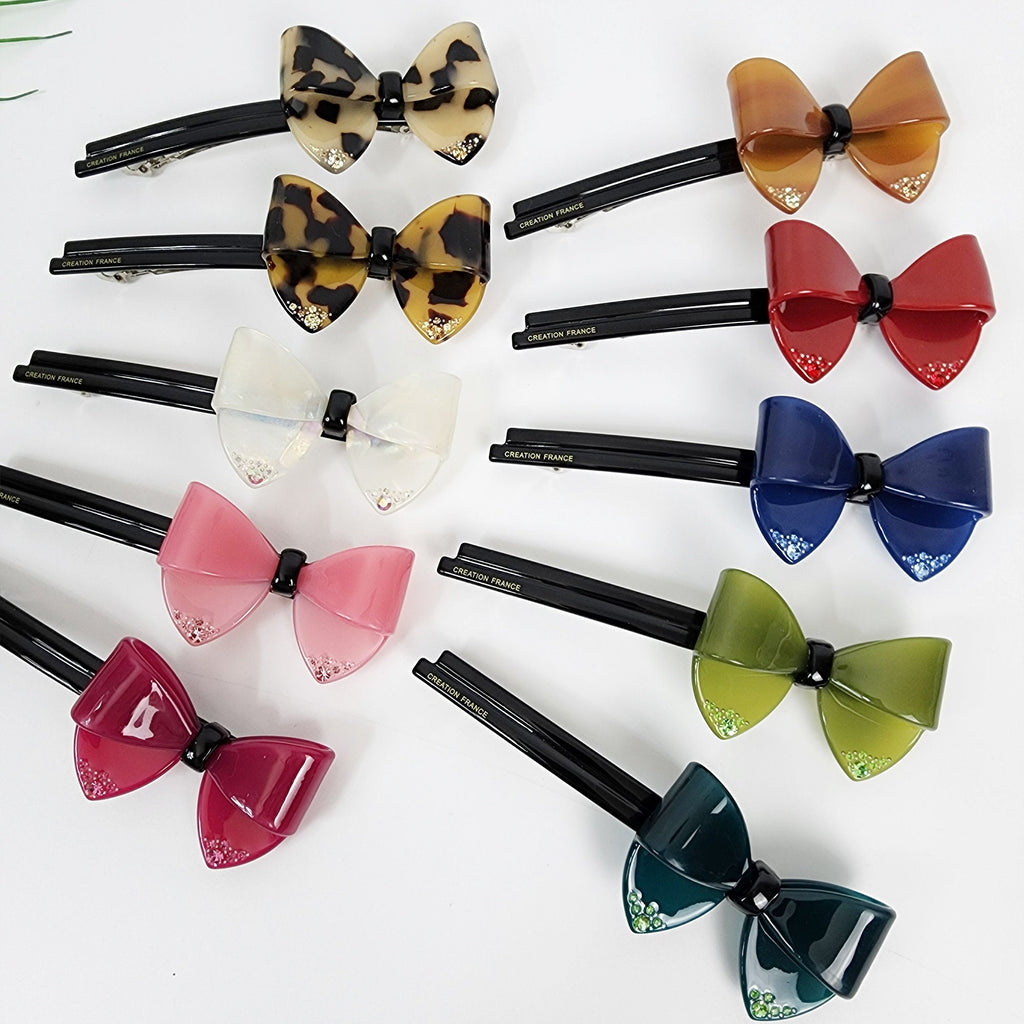 Cellulose acetate large bow barrette with rhinestone crystal accents in tortoise shell pattern. 5 inch french style hair clip for women holding full ponytail or half up hairstyle. Ten black hair clips with different colored bows. The bows are made of plastic and have a shiny finish. The bows are in the following colors: brown, red, blue, green, white, pink, burgundy, and two shades of green. Each bow has a small rhinestone in the center. The clips are labeled 'Creation France'.