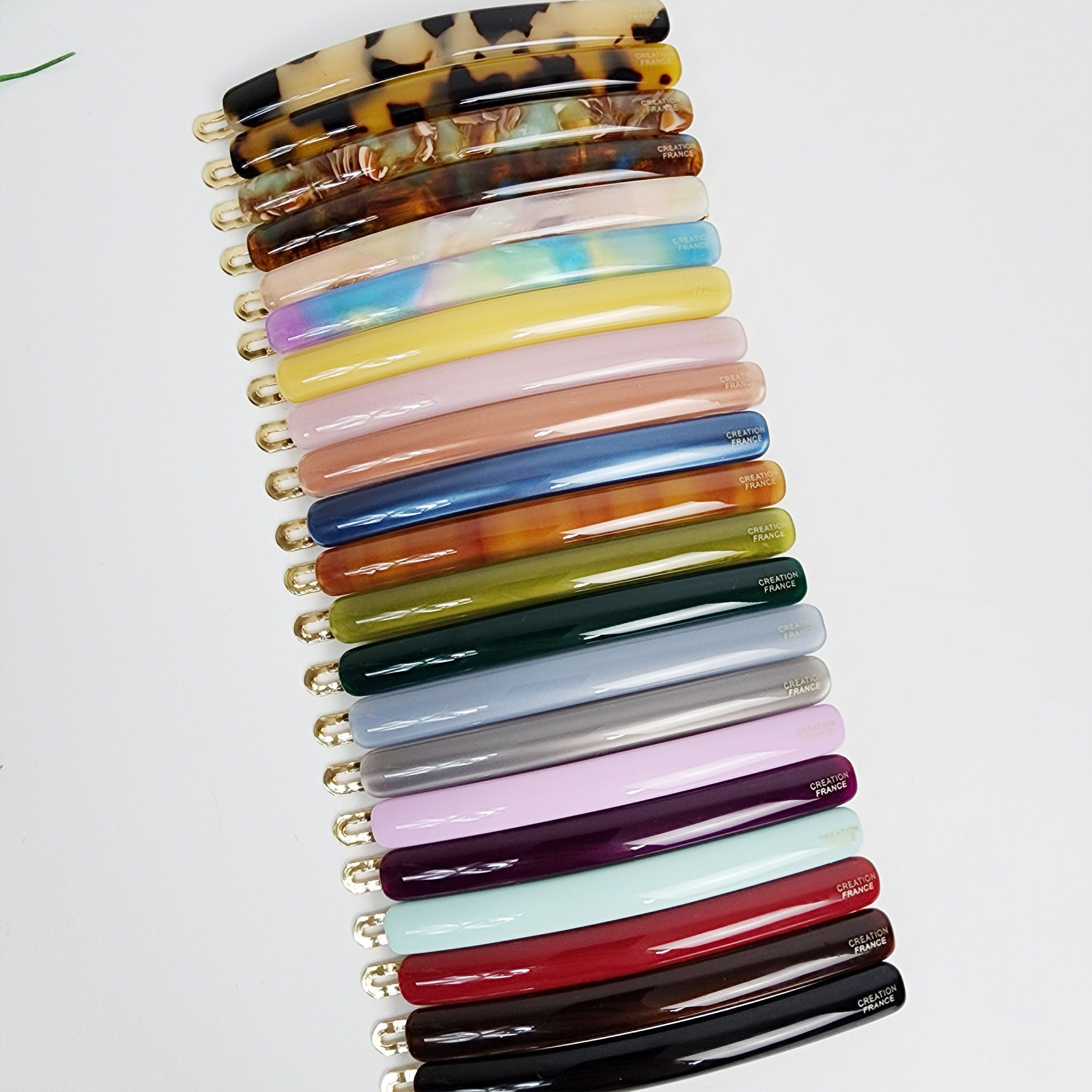 Long cellulose acetate hair barrette in tortoise shell and pastel colors. Minimalist thin hair clip for side bangs or half updo styling. A collection of 18 hair clips in various colors, including black, brown, red, blue, green, and purple. The clips are made of a shiny, smooth material and have a curved shape. Each clip has a gold metal clasp and the words 'CREATION FRANCE' printed on the back.