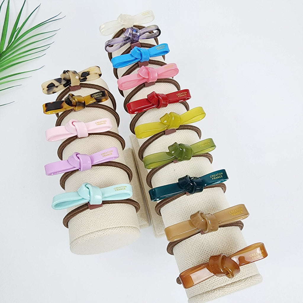 Cellulose acetate mini twist ribbon hair tie in leopard and pastel colors. Small knotted bow ponytail holder. Cute Korean hair accessory. A display of hair clips with bows in various colors including pink, blue, green, red, brown, and yellow. The hair clips are arranged on a white display stand with the brand name 'CREATION FRANCE' printed on the clips.