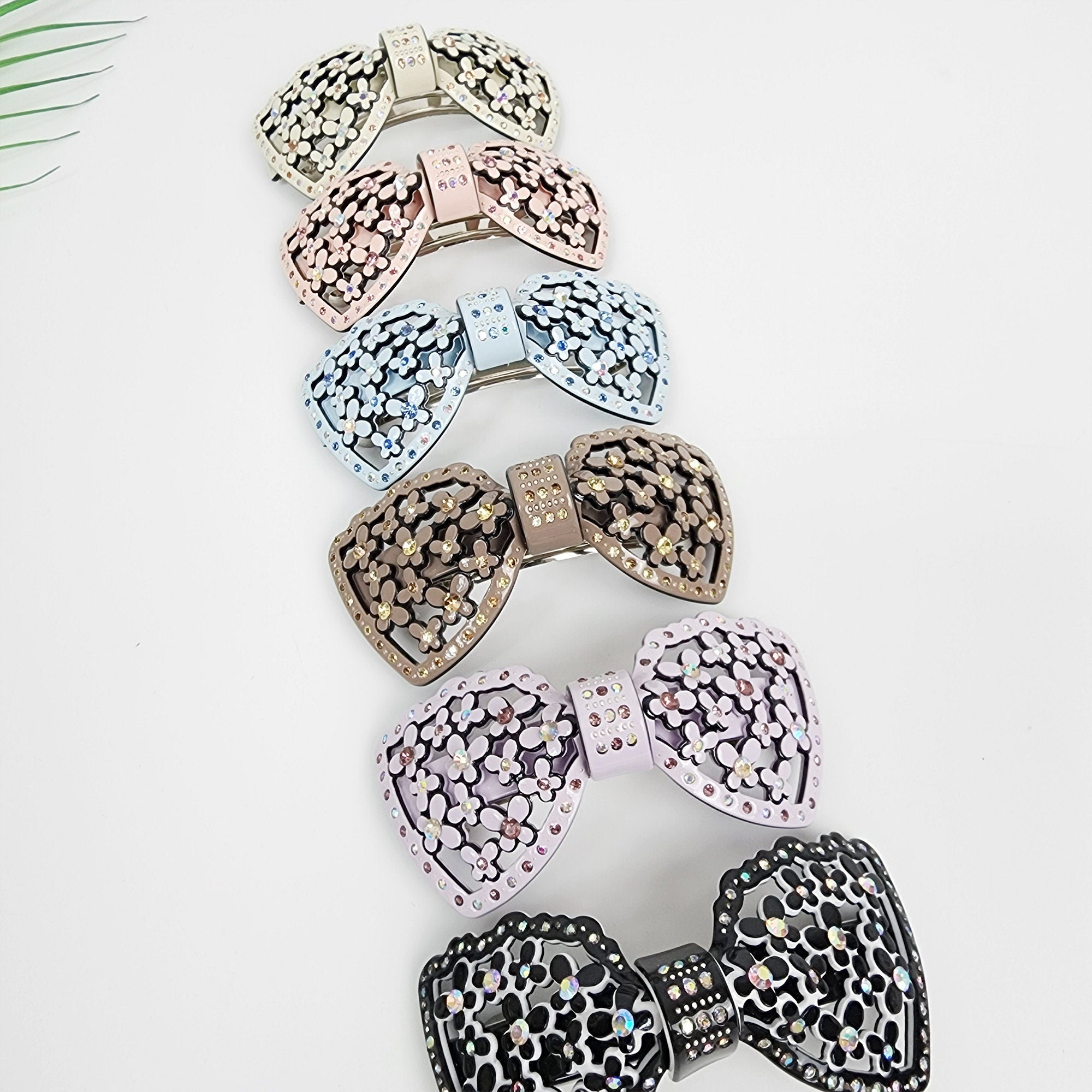 Pastel acetate bow barrette with rhinestone flower pattern and cutout design. Elegant 3.9 inch floral hair clip for thick hair, available in pink, blue, and black. Sparkly hair accessory for women. Six hair clips in the shape of a bow with rhinestones. The bows are in different colors, including light pink, light blue, brown, lavender, and black.