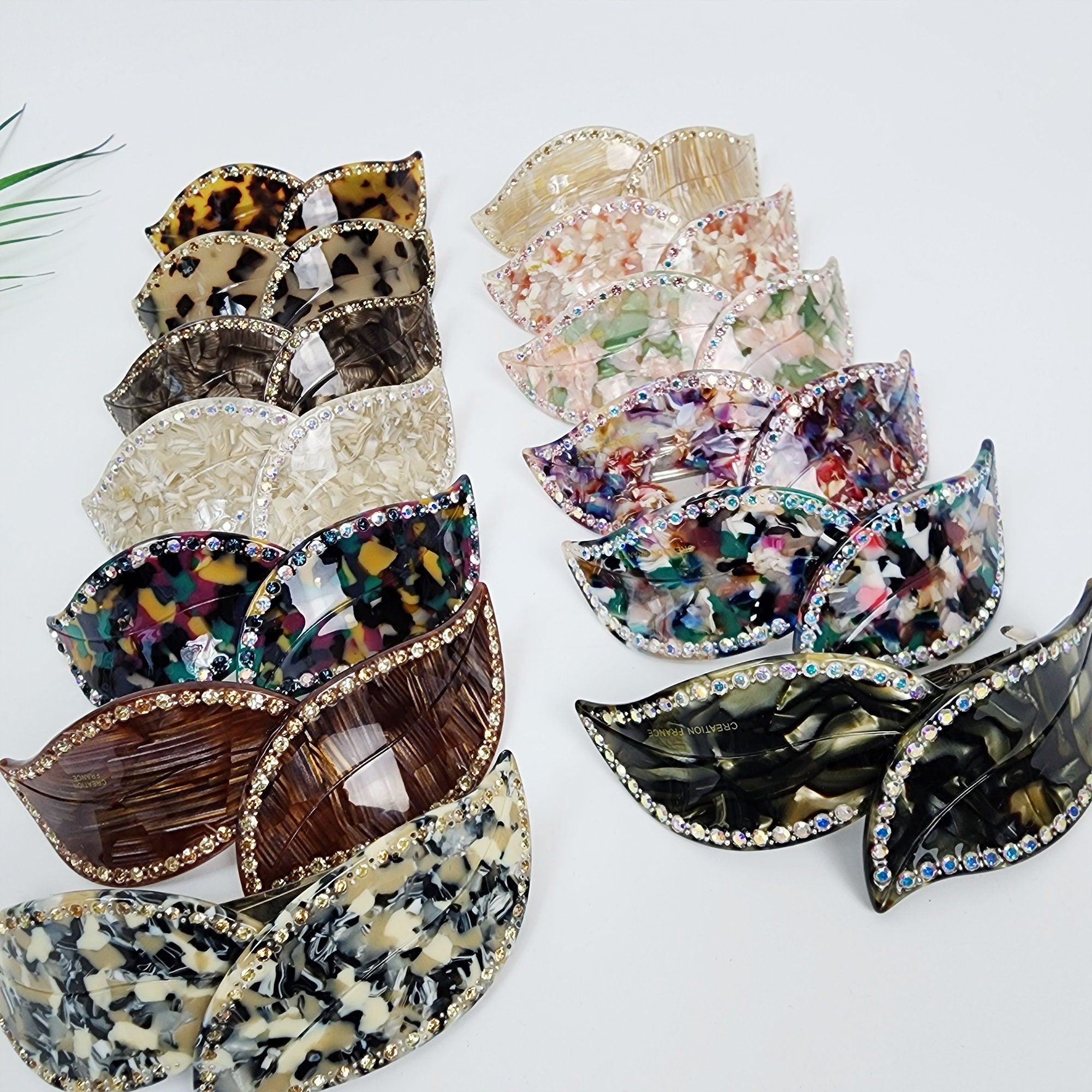 Cellulose acetate leaf shaped hair barrette with crystal rhinestone border. Large 4.1 inch automatic hair clip featuring tortoise and marble patterns for half-up hairstyles. A collection of twelve pairs of leaf-shaped hair clips. The clips are made of clear, brown, black, and multi-colored plastic. Each clip has a row of rhinestones around the edge. The clips are arranged in a stack, with the top clip being a pair of black clips with the words 'Caution on Hair' printed on the back.