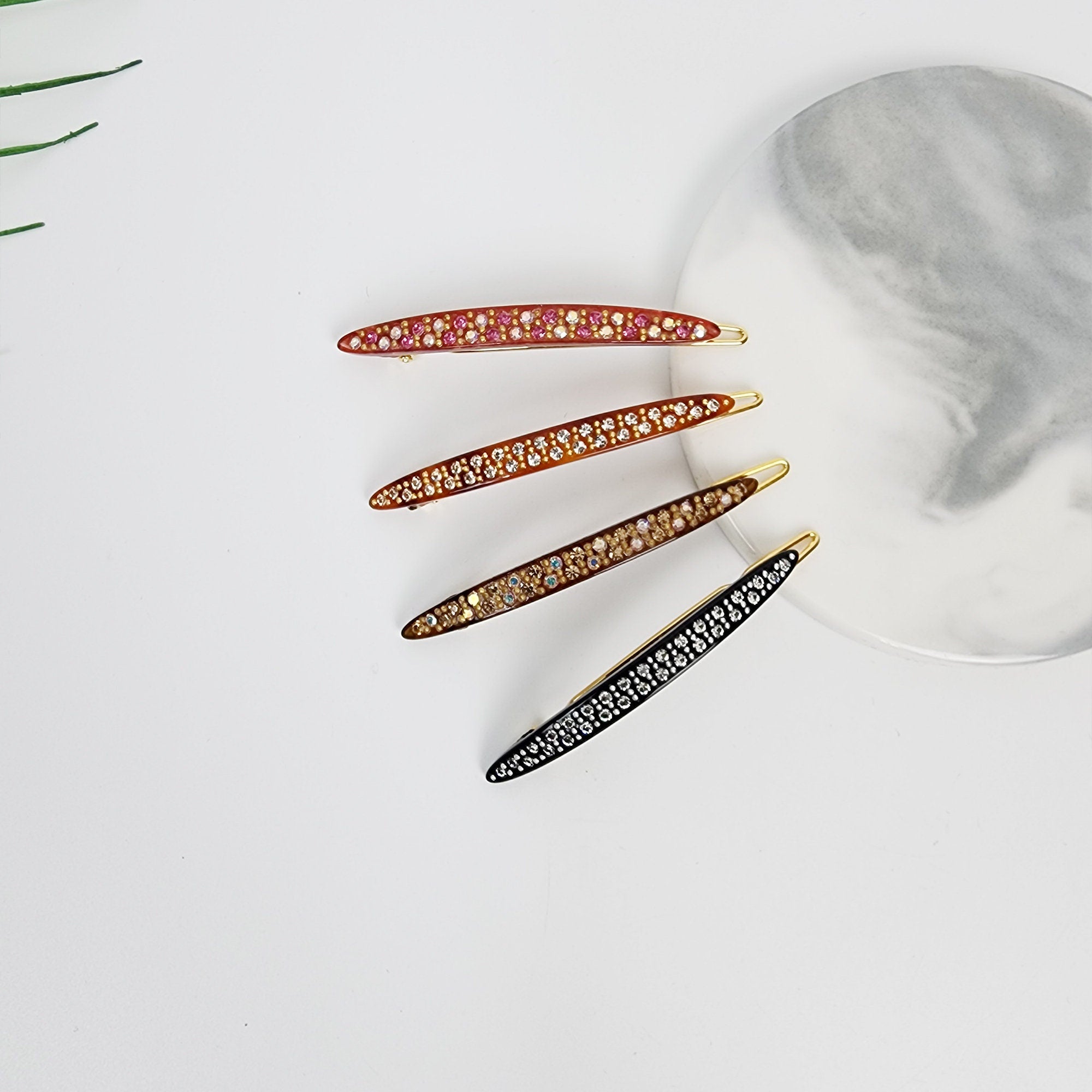 Cellulose Acetate Slim Snap Clip with Crystal Embellishments Long Oval Barrette for Bangs Tortoise Shell Pattern Korean Hair Accessory Four jeweled hair clips in varying colors. The clips are curved and feature a line of small, sparkling stones. Colors include red, brown, gold, and black. They are arranged on a white surface with a marble-patterned plate.