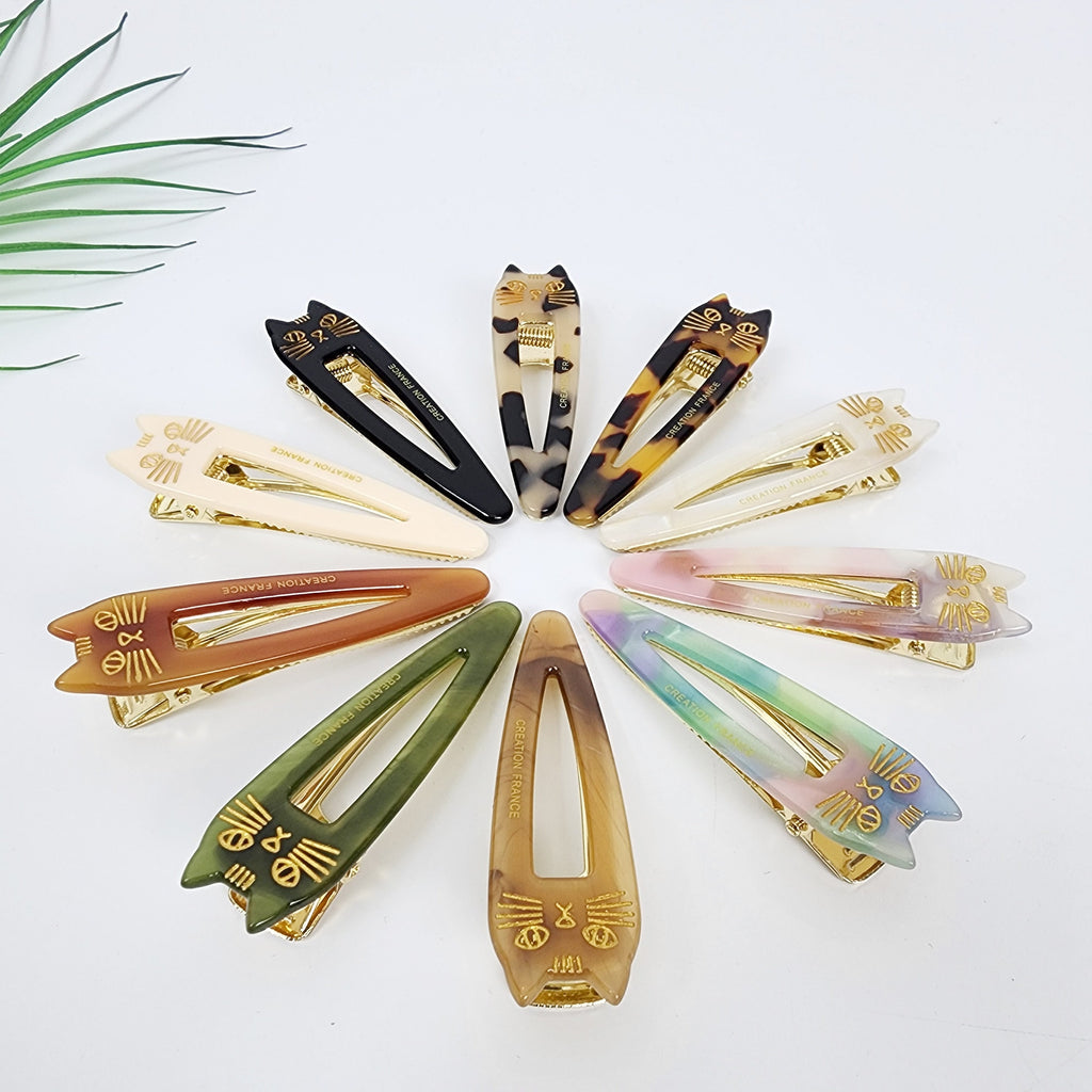Cat shaped cellulose acetate hair slide with gold whiskers details. Tortoise and marble pattern alligator clip for side bangs, kawaii hair accessory. Ten cat-shaped hair clips in various colors, including black, brown, beige, green, pink, and purple. Each clip has a gold metal clasp and a gold cat face design with the words 'CREATION FRANCE' printed on the back.
