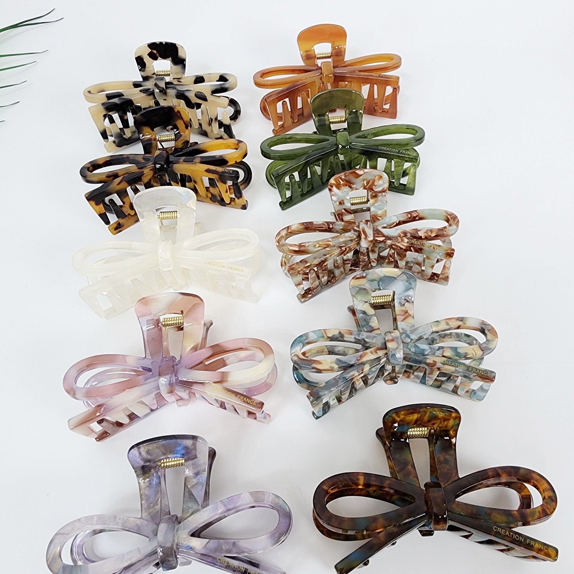 Cellulose acetate hair claw in a looped ribbon shape. 3.1 inch medium jaw clip in tortoise shell and solid colors for half-up hairstyles. Ten different hair clips with a bow design. The clips are made of plastic and have a marbled or tortoiseshell pattern. The clips are in various colors, including brown, green, white, pink, and blue. The clips have the words 'CREATION FRANCE' printed on them.
