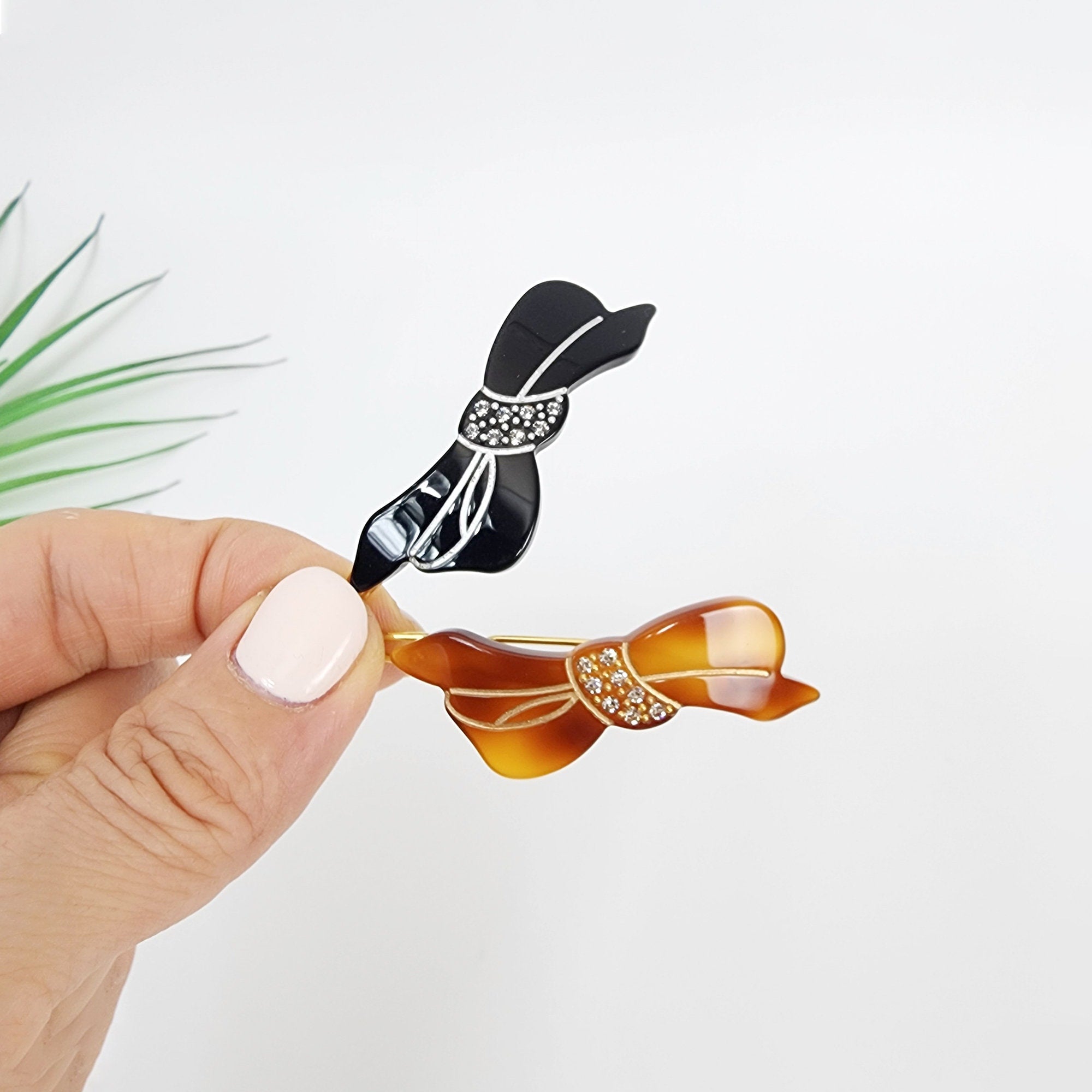 Small cellulose acetate ribbon hair clip with rhinestone accents. Mini bow barrette in tortoise and black for side bangs styling. Two hair clips in the shape of bows. One is black with white rhinestones and the other is brown with clear rhinestones.