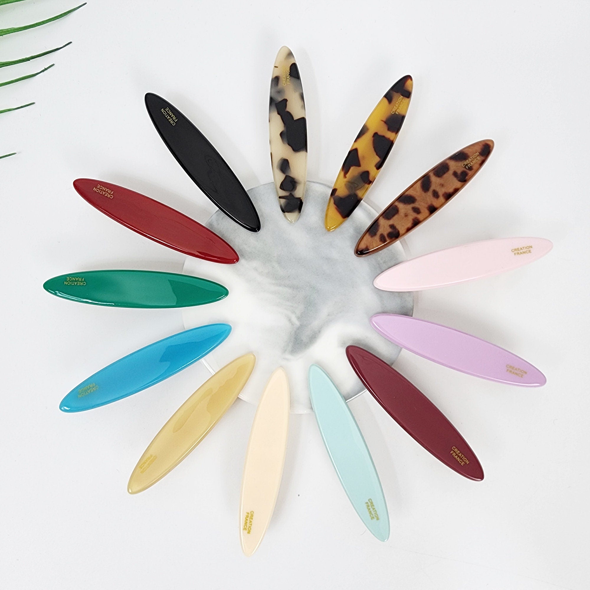 Cellulose acetate oval alligator clip in tortoise shell and pastel tones. Flat 2.8 inch side hair barrette for holding bangs and fine hair securely.  A collection of twelve oval-shaped hair clips in various colors, including black, red, green, blue, yellow, white, pink, purple, and brown. The clips are arranged in a circle on a white marble surface. Each clip has the words 'CREATIVE FRANCE' printed in gold lettering.