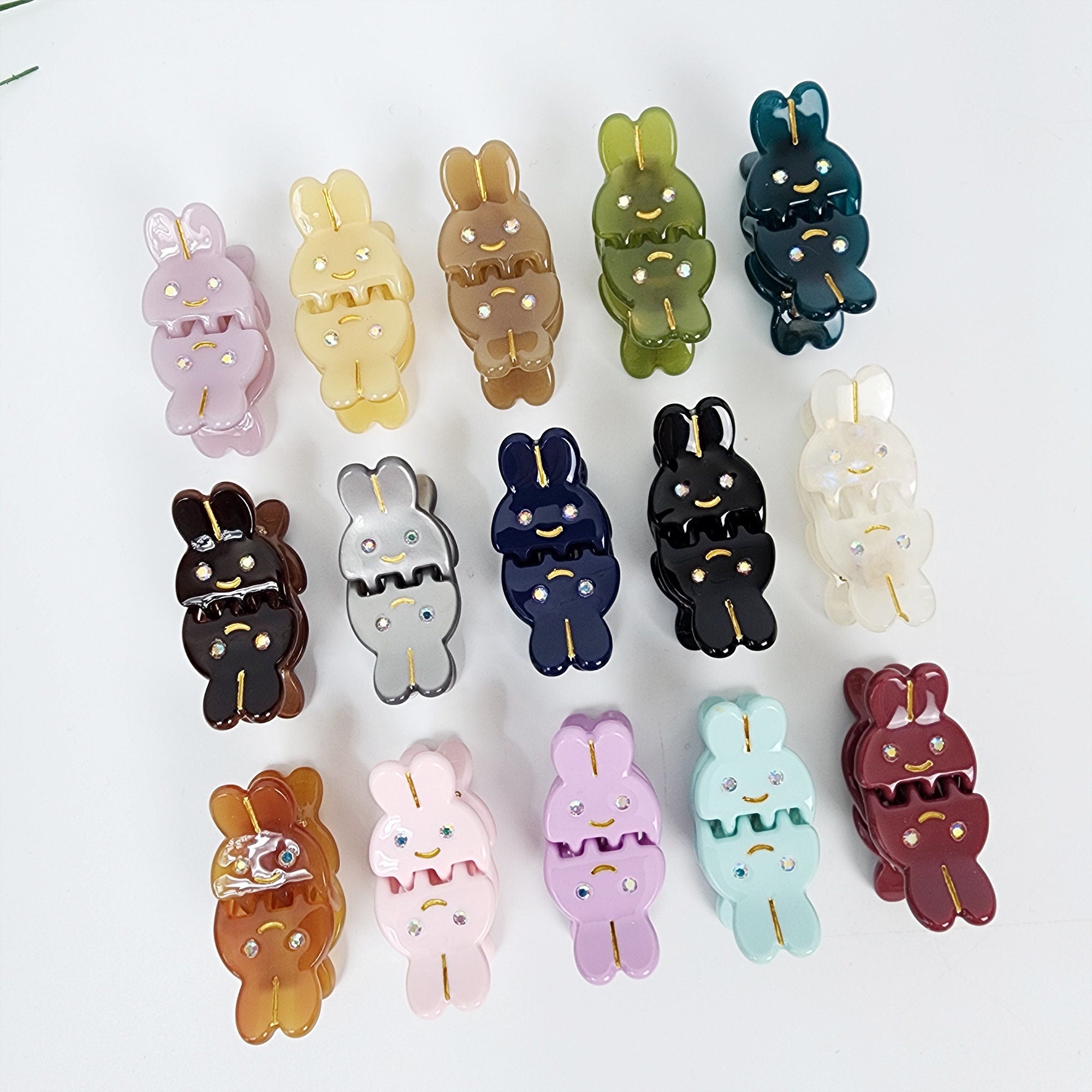 Cellulose acetate mini bunny hair claw clips set of 2 cute rabbit design with rhinestone accents small jaw clips for girls. A collection of 15 colorful plastic bunny hair clips. The clips are in various colors including pink, purple, blue, green, brown, black, and white. Each clip has a small, sparkly rhinestone on the front.