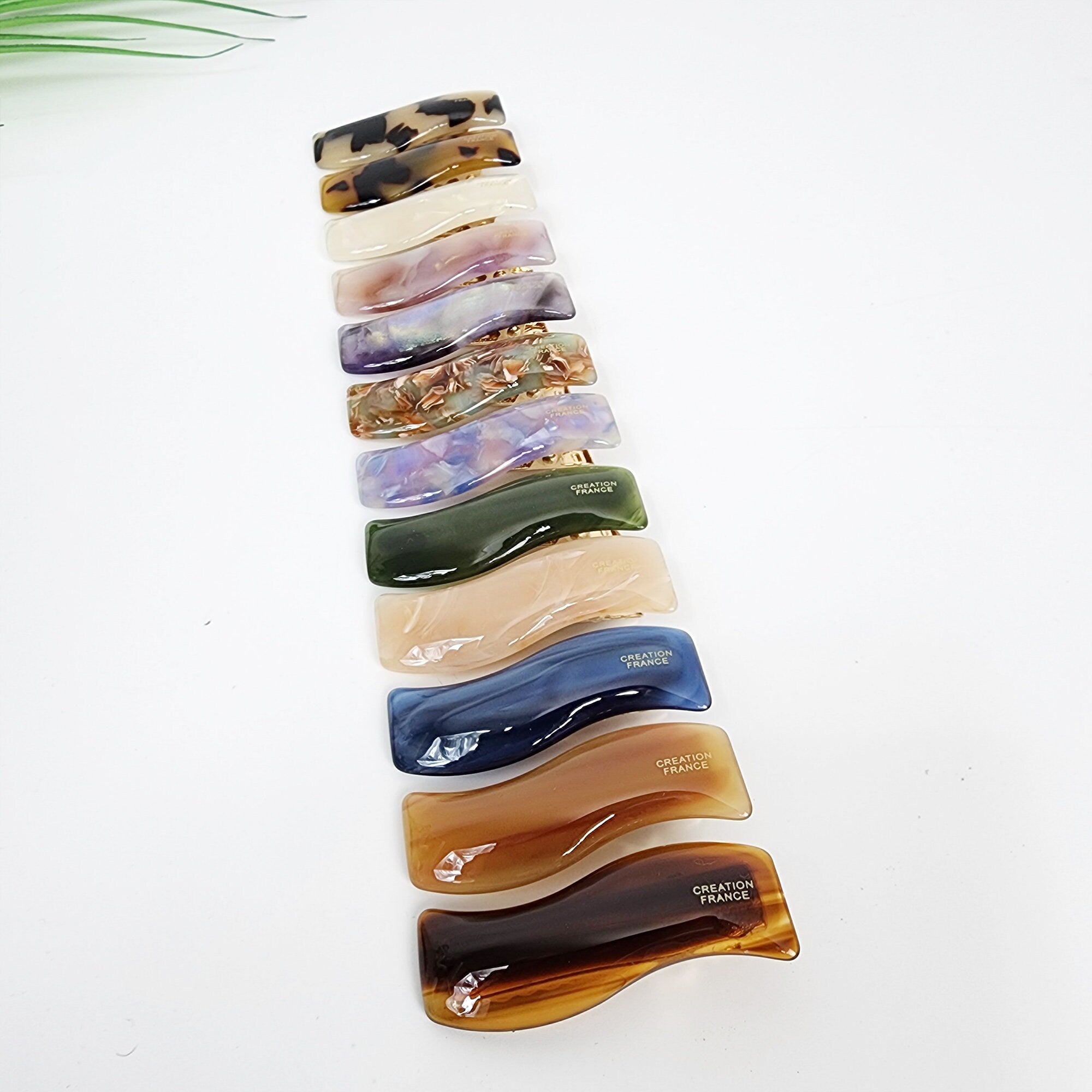 Small cellulose acetate wavy hair clip in tortoise shell and marble patterns, featuring a 1.9 inch squiggle design alligator clip for holding bangs and fine side hair sections.  A set of 12 hair clips in various colors and patterns. The clips are all made of plastic and have a curved shape. The clips are arranged in a row from top to bottom, with the top clip being a brown and black patterned clip. The clips are all labeled with the words 'CREATION FRANCE'.