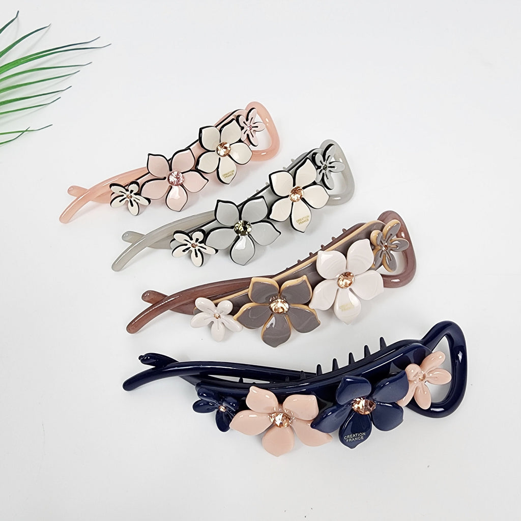 Cellulose acetate banana clip featuring pastel flowers with crystal centers. 4.3 inch twisted hair accessory perfect for romantic half-up hairstyles. Four hair clips with floral designs. The clips are in different colors, including pink, gray, brown, and blue. Each clip has a comb-like design on one side and a floral design on the other side. The floral designs are made of plastic and have rhinestones in the center of each flower. The blue clip has the words 'Creation France' on the side