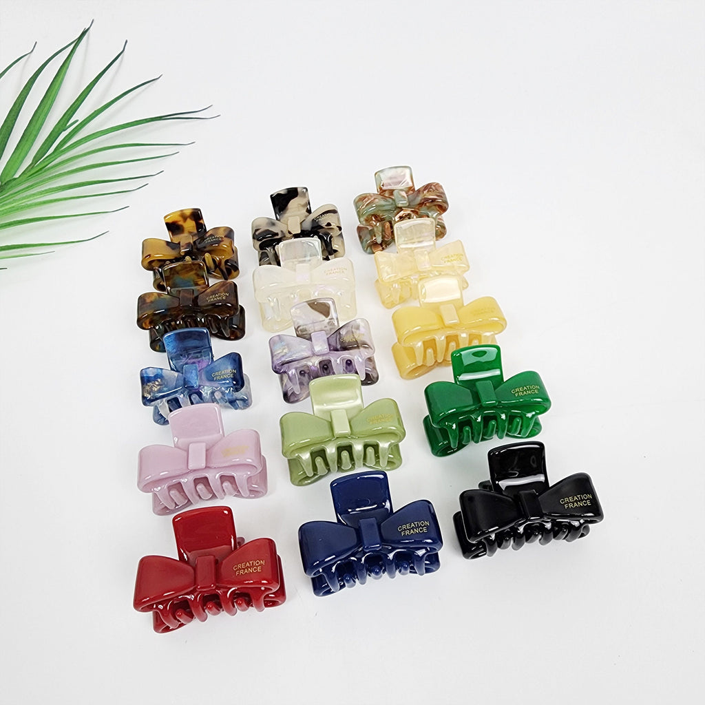 Small cellulose acetate bow hair claw clip featuring tortoise and pastel colors. 1.4 inch mini jaw clip perfect for side bangs styling. A collection of 14 hair clips with a bow design. The clips are made of plastic and come in a variety of colors including brown, white, yellow, purple, green, pink, red, blue, and black. Each clip has the words 'CREATION FRANCE' printed on it in gold lettering.