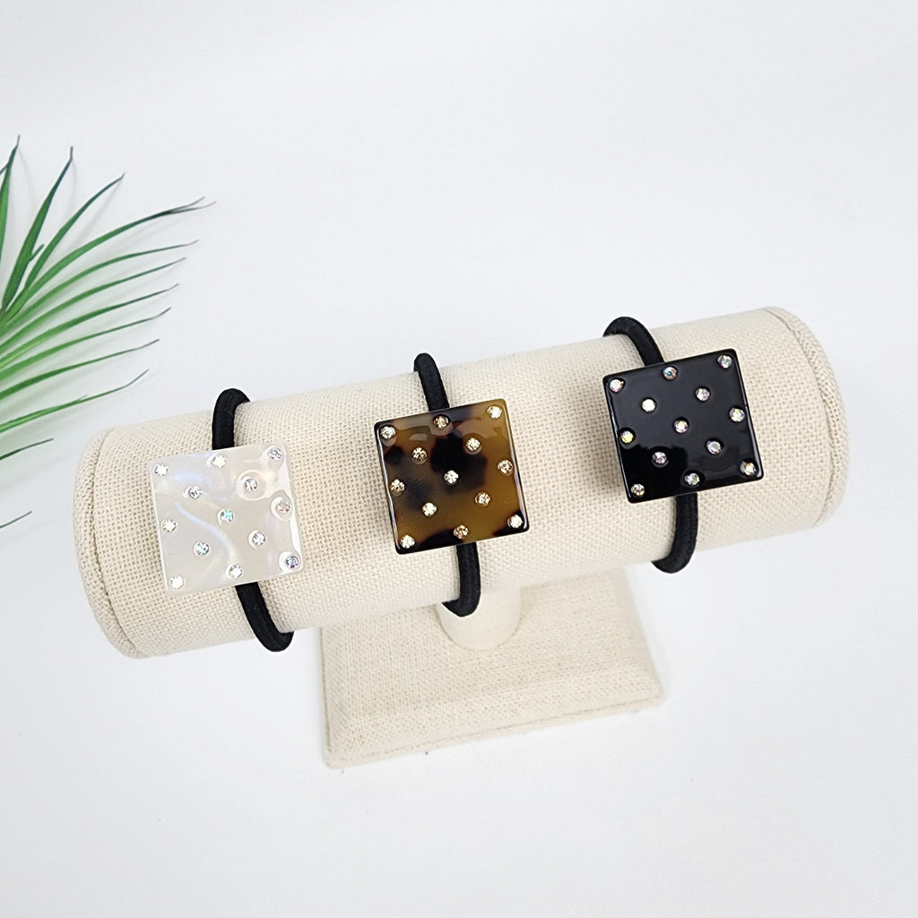 Cellulose acetate square hair tie with rhinestones, geometric ponytail holder with crystals, minimalist tortoise shell accessory for birthday gifts. Three hair scrunchies with square-shaped embellishments. The embellishments are made of a tortoise shell-like material with rhinestones. The scrunchies are displayed on a white fabric stand.